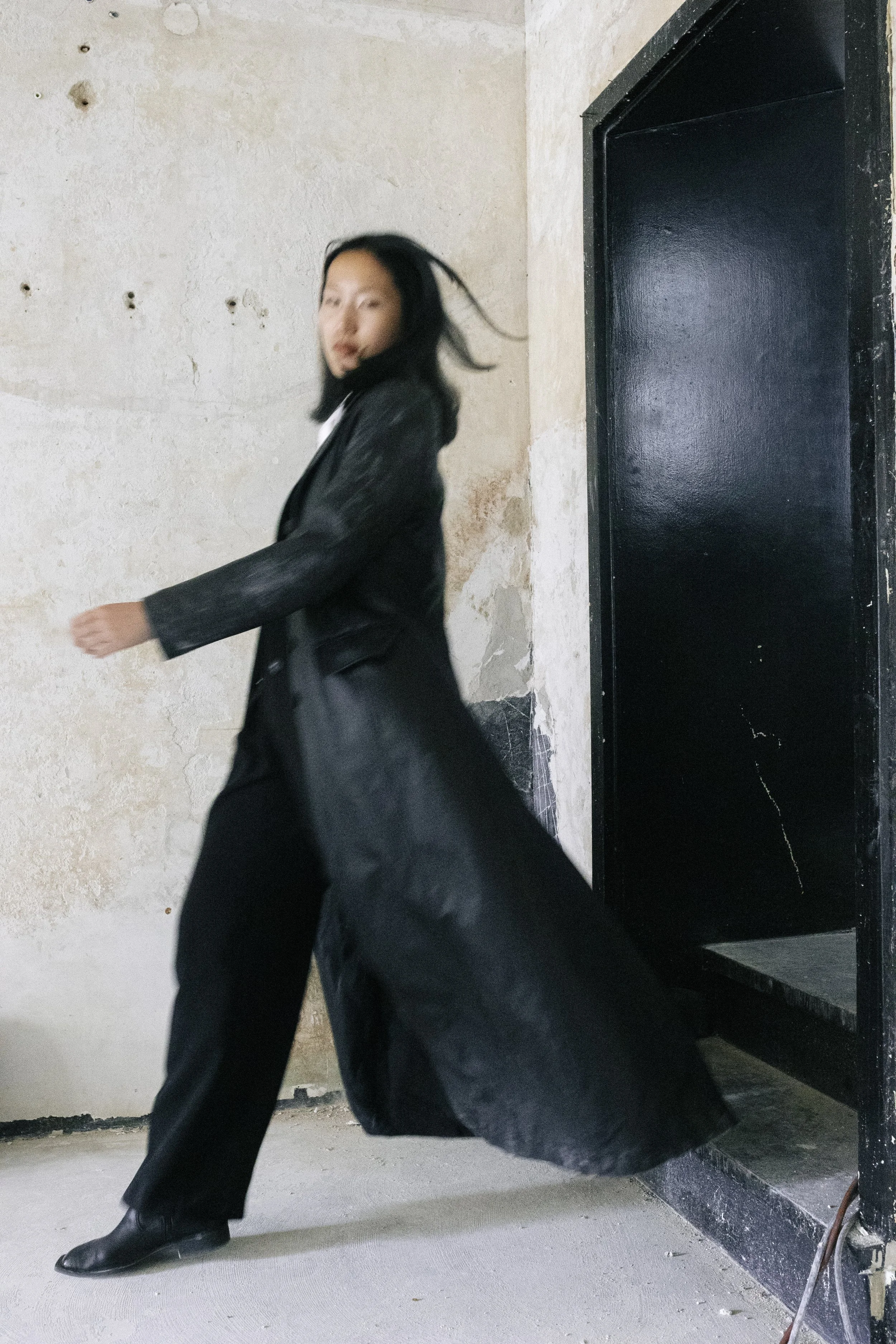 Eine Frau in schwarzer Kleidung läuft an der Kamera vorbei in einem alten, leicht beschädigten Raum mit einer schwarzen Tür.