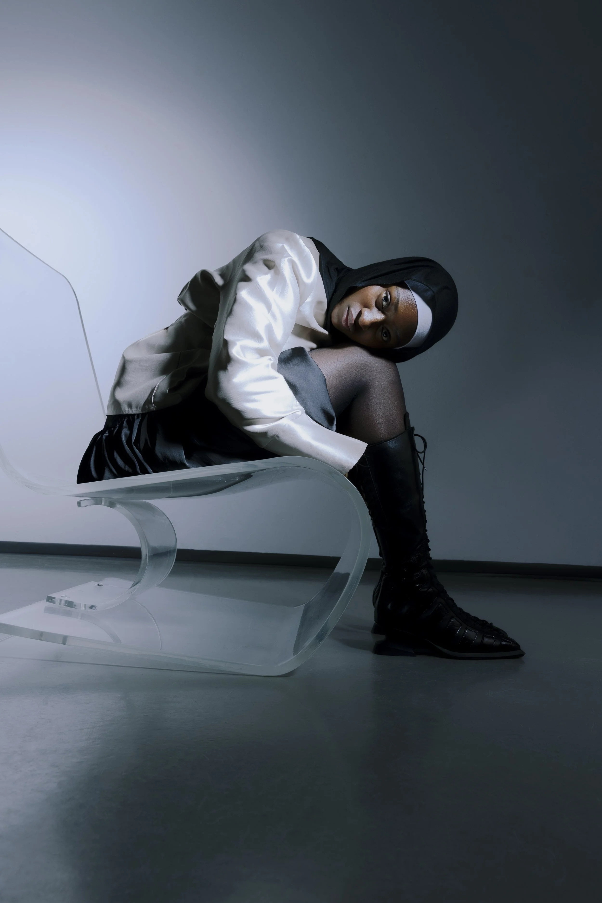 Junge Frau in modischer Kleidung sitzt auf einem transparenten Sessel, in einem dunklen Raum, Blick wendet sich nach oben, schwarz-weißer Hoodies, schwarze Strümpfe und Stiefel.