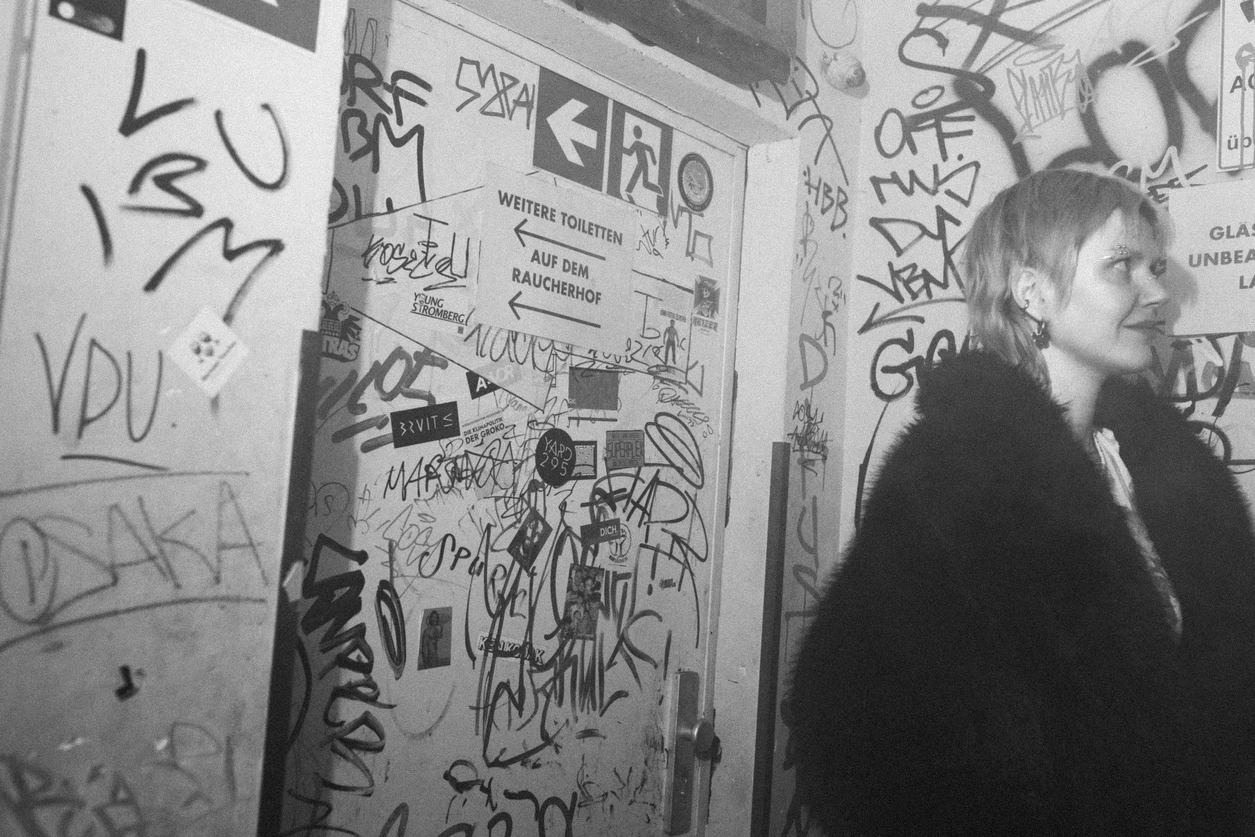 Eine Frau mit kurzen Haaren und einem schwarzen Pelzmantel steht in einem graffitiüberdeckten Raum, schwarz weiß Aufnahme.