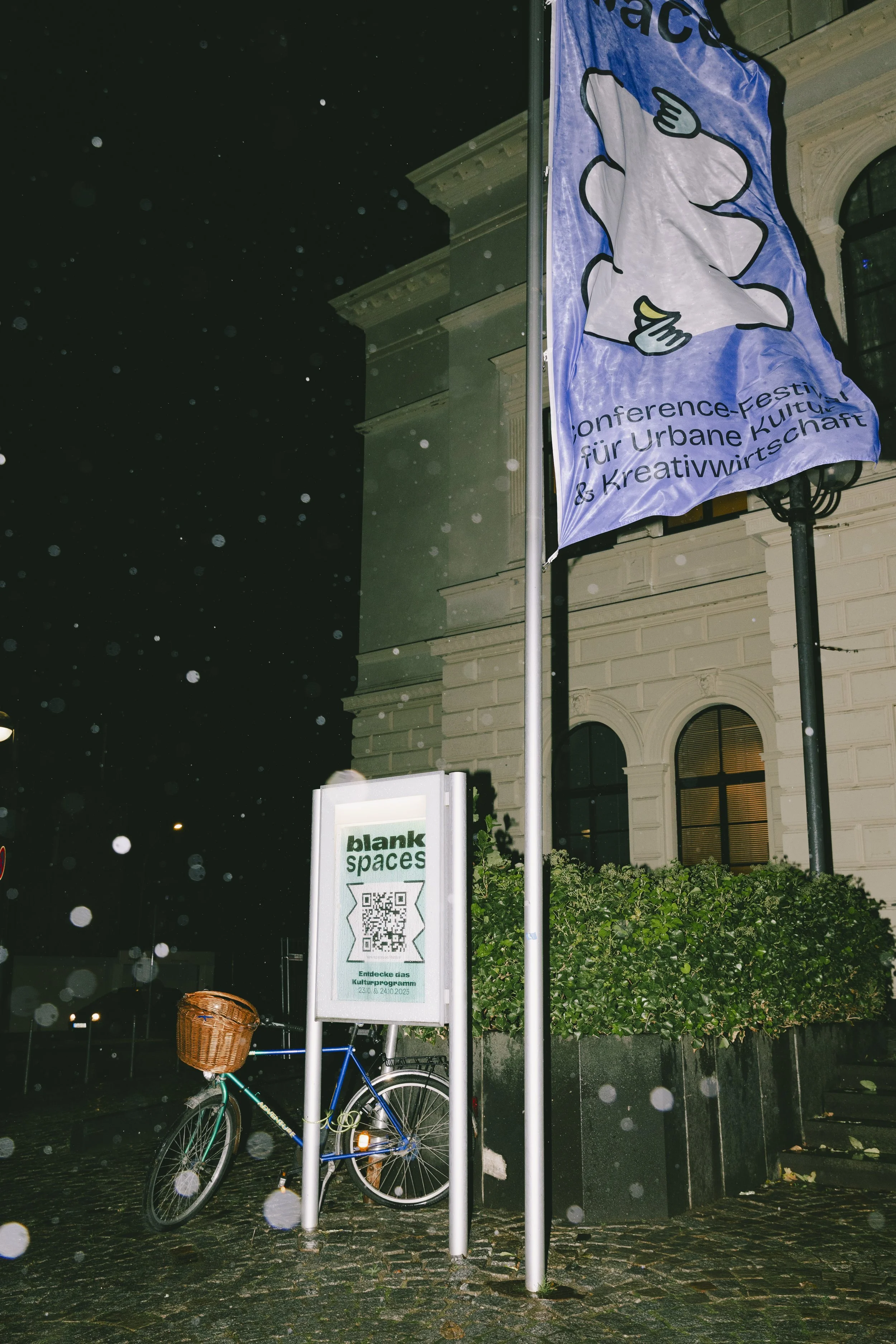 Nachtaufnahme eines alten Gebäudes mit einem Fahrrad mit Korb davor und einer lila Fahne die im Wind weh, darauf Werbung für ein Festival.