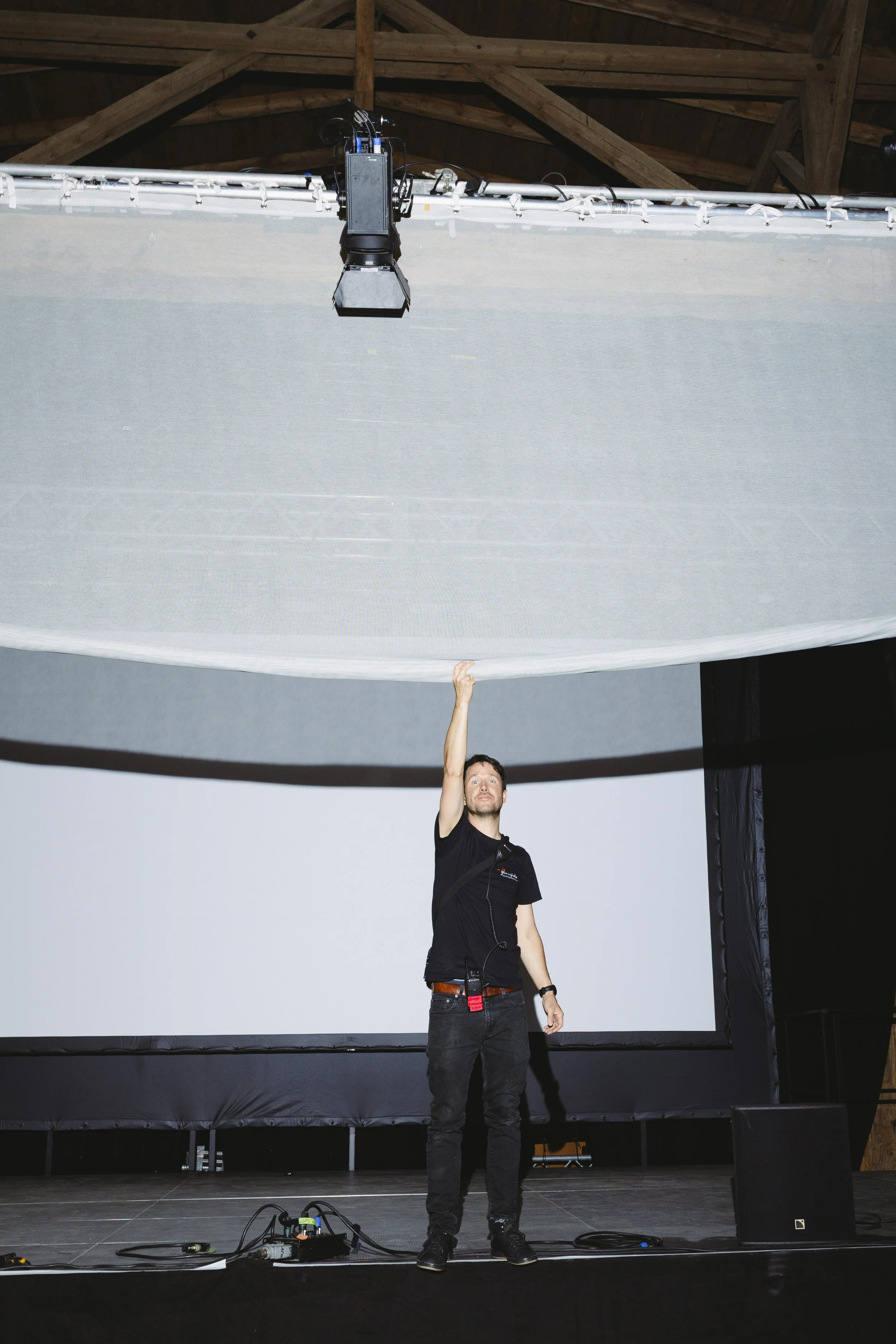 Ein Mann steht auf einer Bühne vor einer großen Leinwand, hebt die Hand und zeigt nach oben. Über ihm hängt eine große graue Projektionsleinwand und oben ist ein technisches Gerät angebracht.