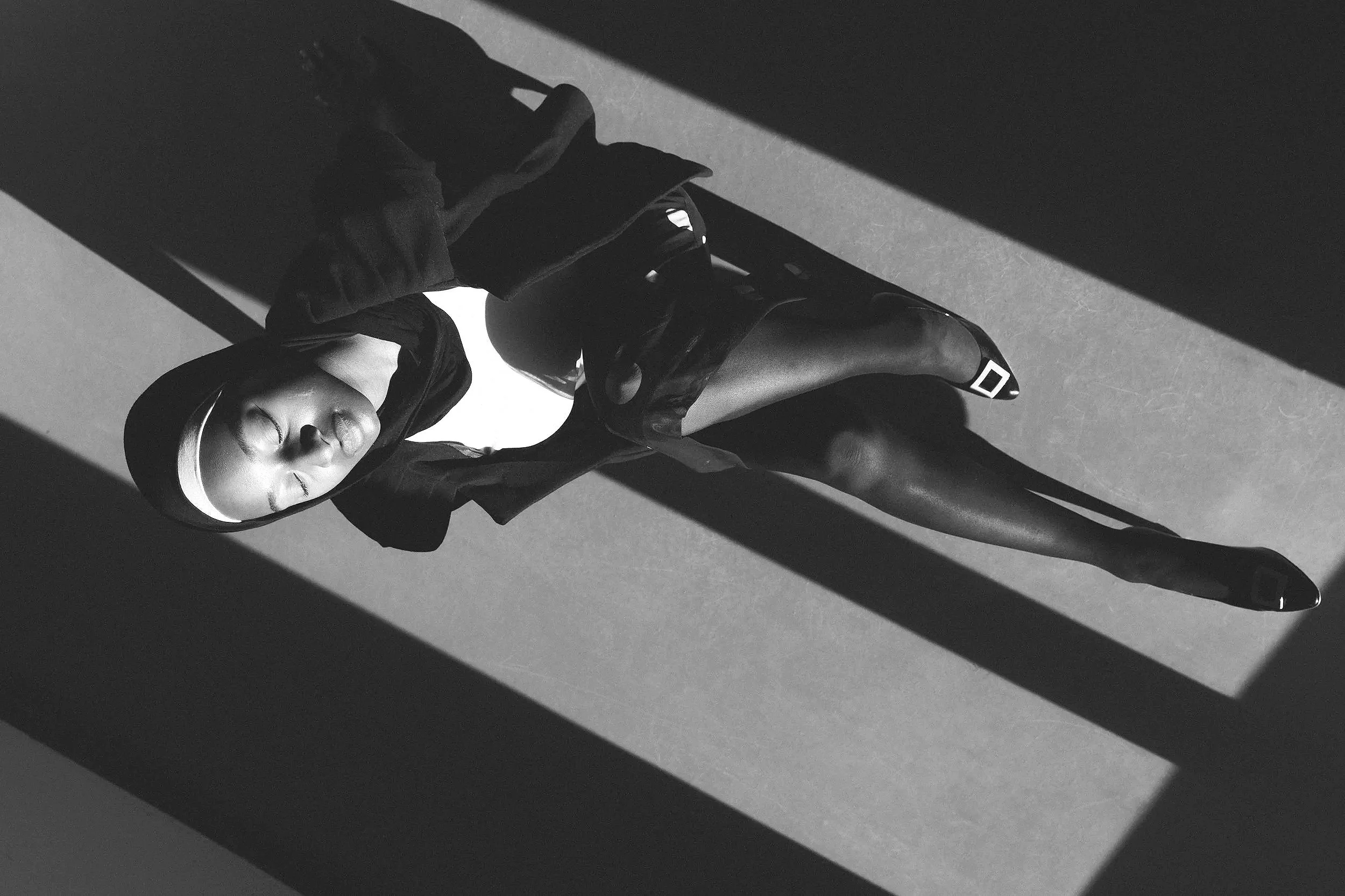 Schwarz-weiße Fotografie eines Mädchens in Sportkleidung, liegend auf dem Boden, mit Blick nach oben, Schattenwürfe auf der Wand hinter ihr.