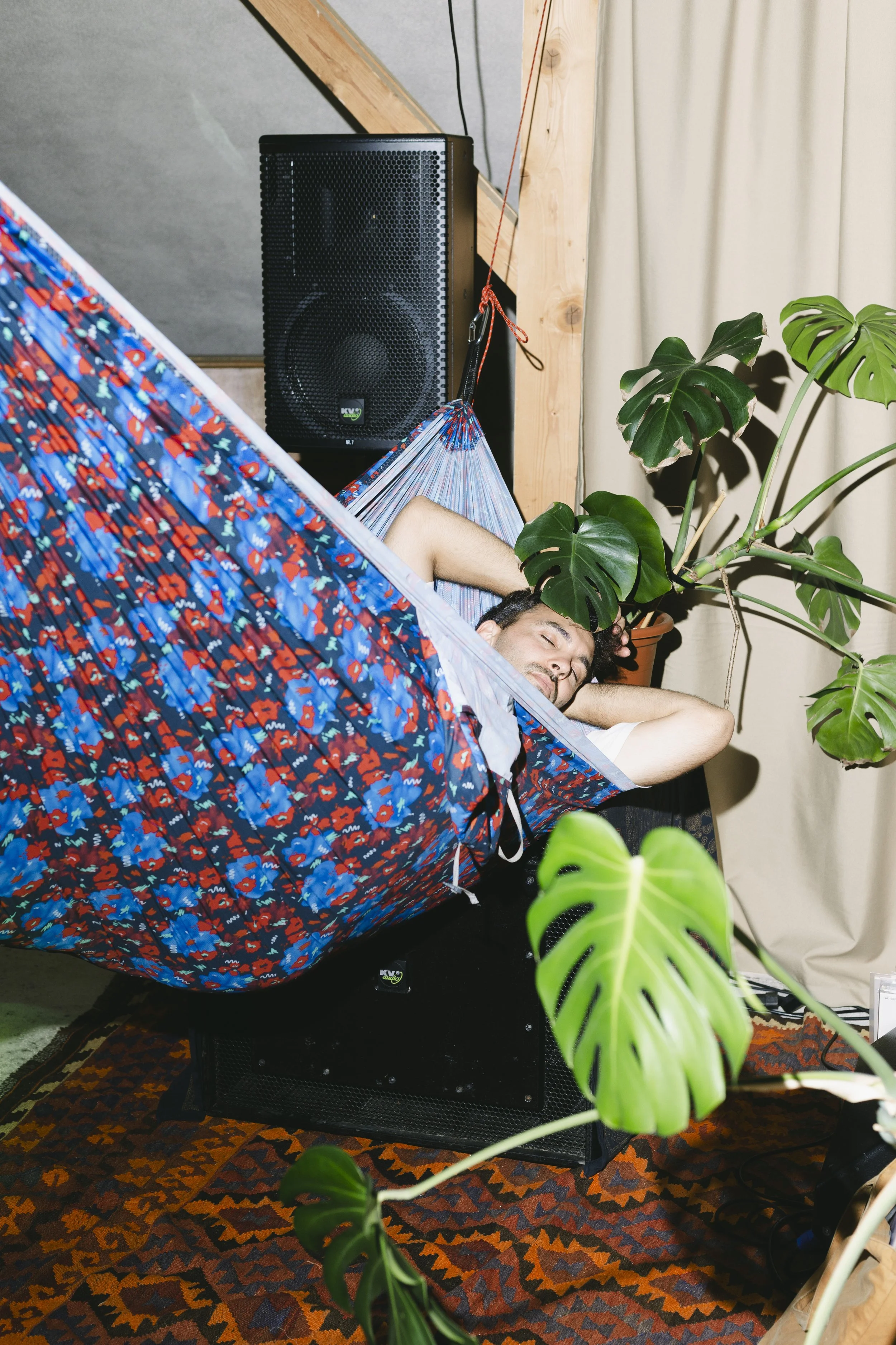 Ein Mann liegt entspannt in einer Hängematte in einem Raum mit Pflanzen und einem Lautsprecher im Hintergrund.