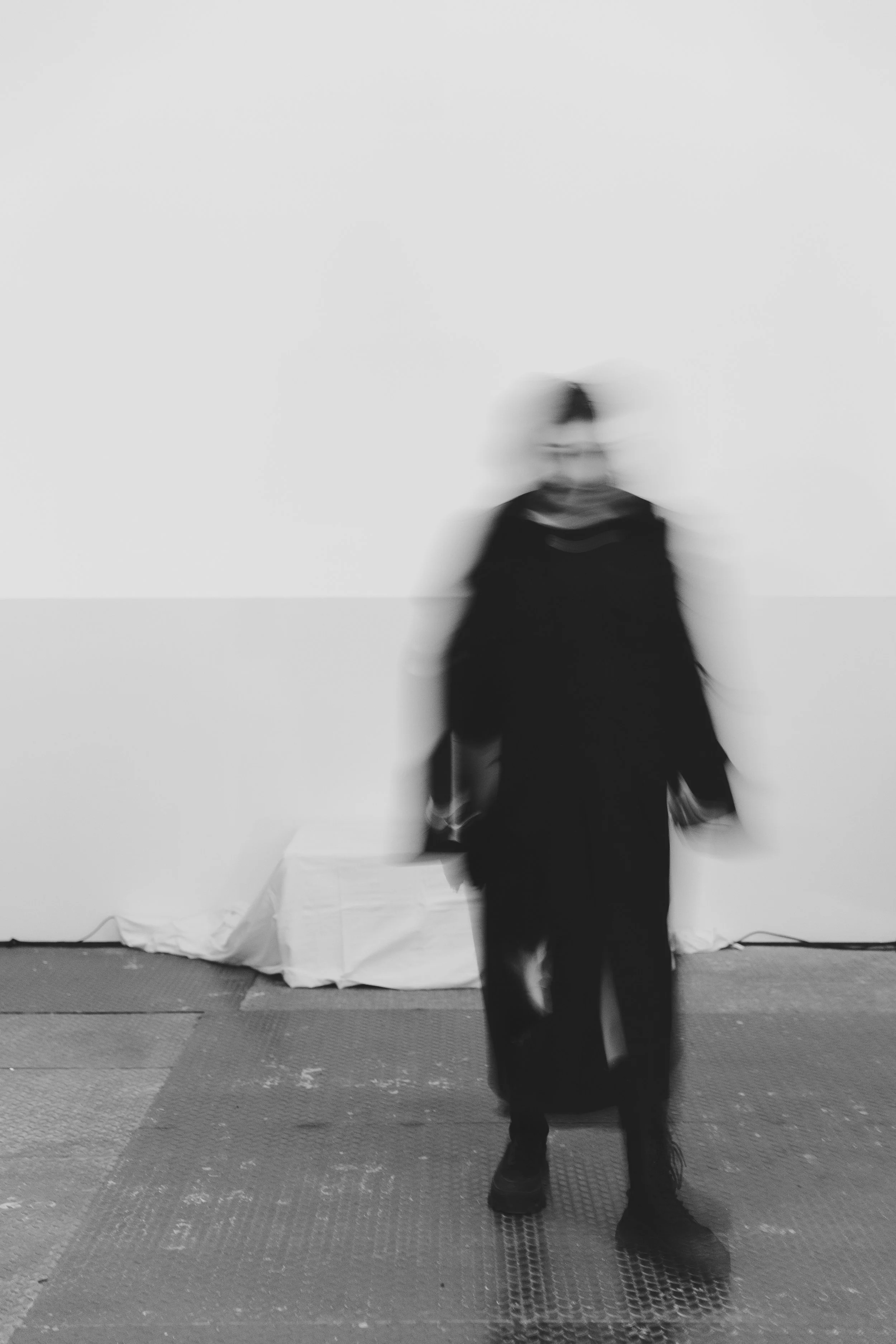 Schwarz-weiß-Foto einer verschwommenen Person vor neutraler Wand. Minimalistische Aufnahme mit Bewegungsunschärfe.