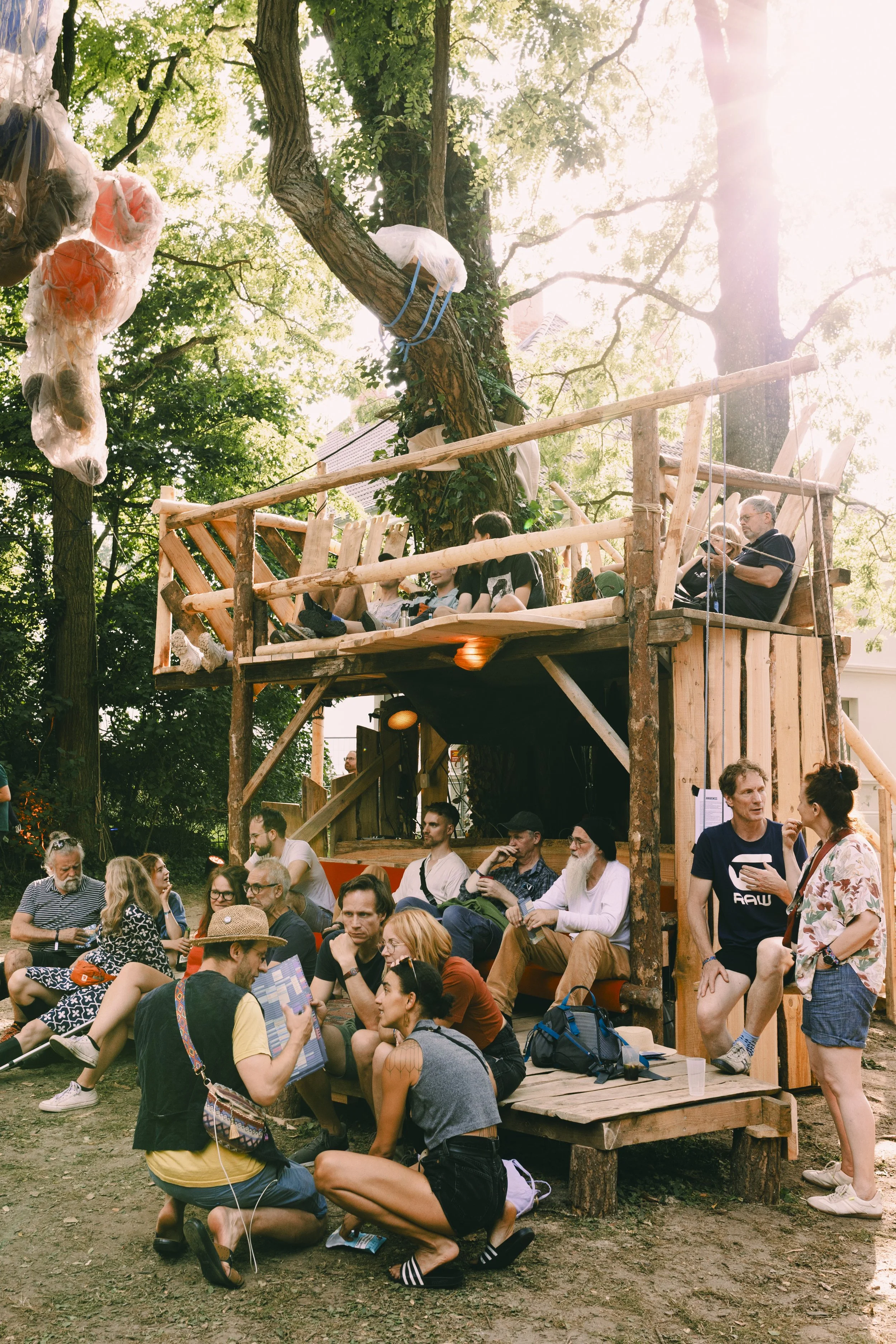 Gruppe von Menschen im Park neben einem Baumhaus, das aus Holz gebaut ist, mit mehreren Ebenen und Sitzbereichen, umgeben von Bäumen und Sonnenlicht.