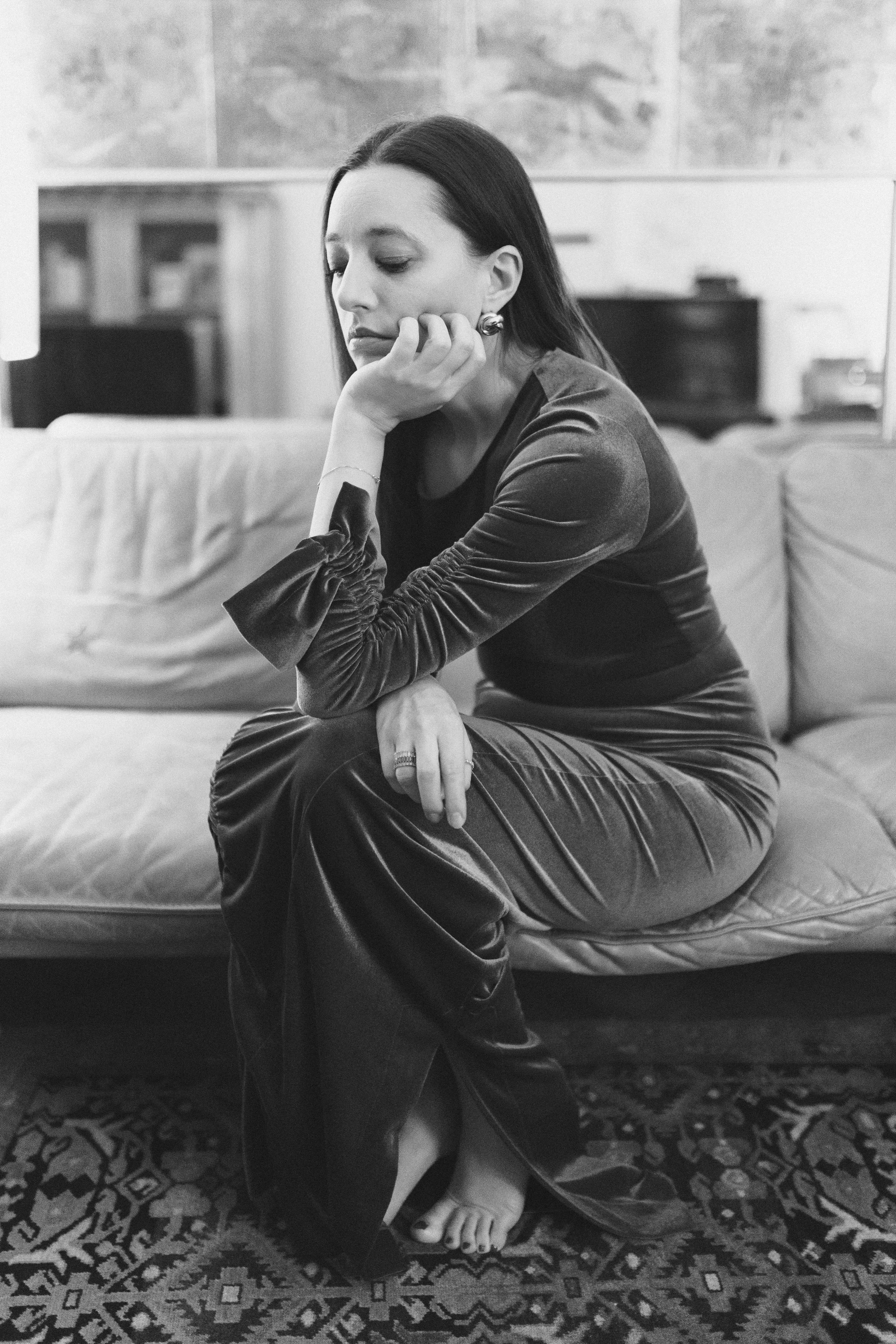 Schwarz-weiß-Fotografie einer Frau, die nachdenklich auf einem Sofa sitzt, mit Hand an ihrem Gesicht, elegante Kleidung und ein Blick nach unten.