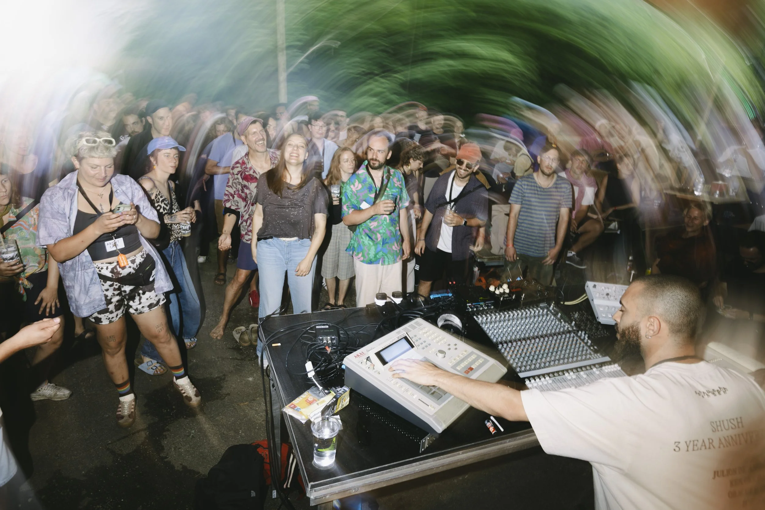 Menschen tanzen und feiern auf einer Party, im Vordergrund ein DJ, der Musik mit einem Mischpult spielt, bei dunklem Licht mit Bewegungsunschärfe und Lichtspuren.