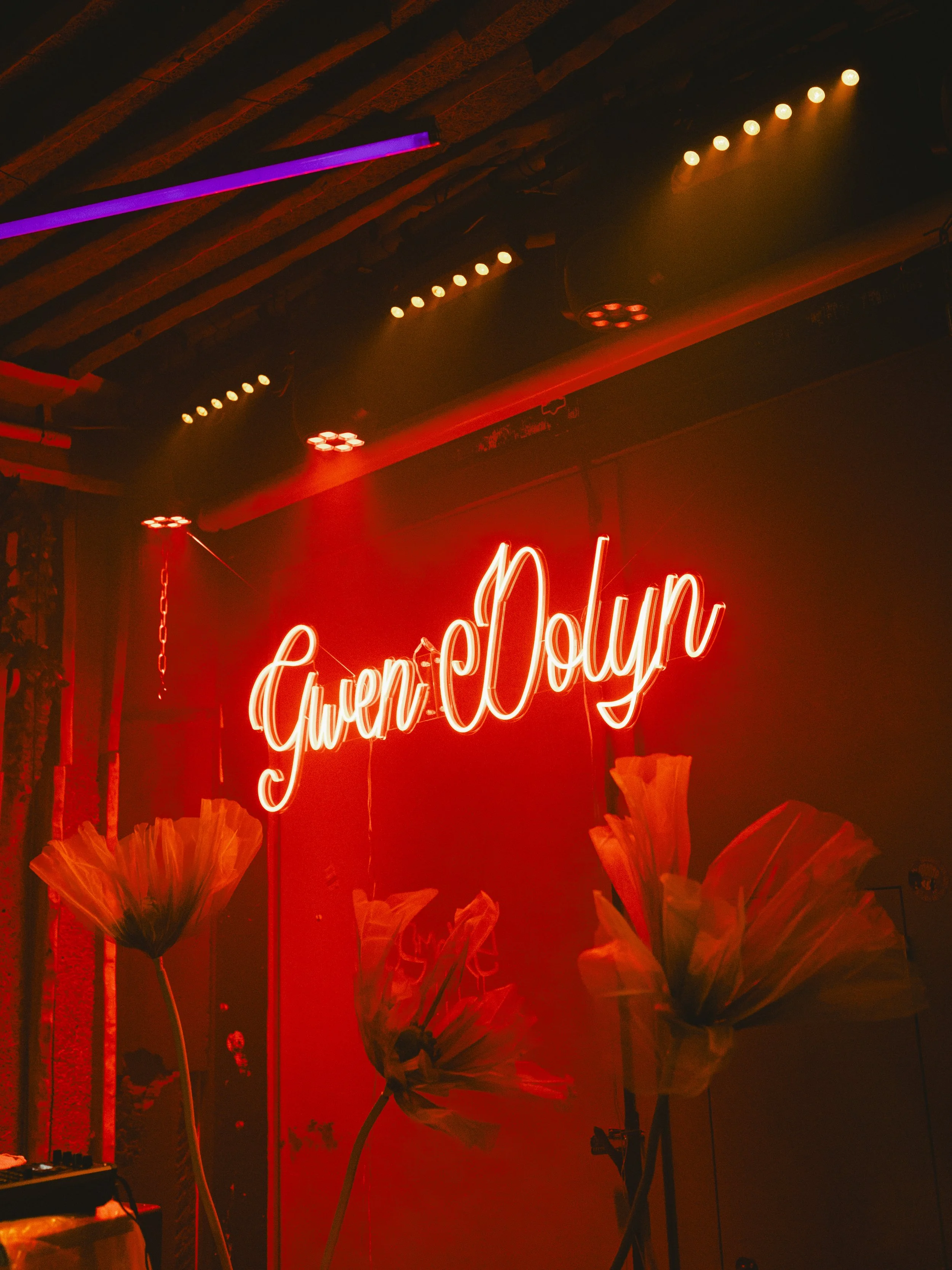 Neon-Schriftzug mit dem Namen 'Gwen-Dolyn' in einem Raum mit roten und blauen Lichteffekten, dekoriert mit großen Stoffblumen.