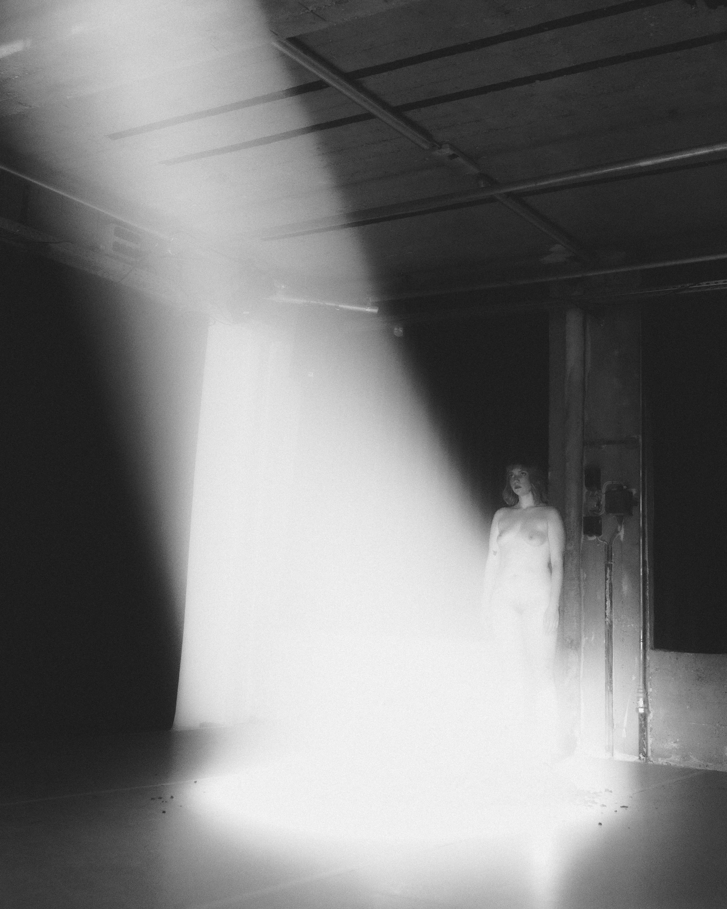 Eine schwarz-weiß Aufnahem einer nackten Performerin in einem dunklen Raum, beleuchtet von einem Strahl hellen Lichts, das aus der oberen linken Ecke kommt. Das Bild ist verschwommen und leicht unscharf.