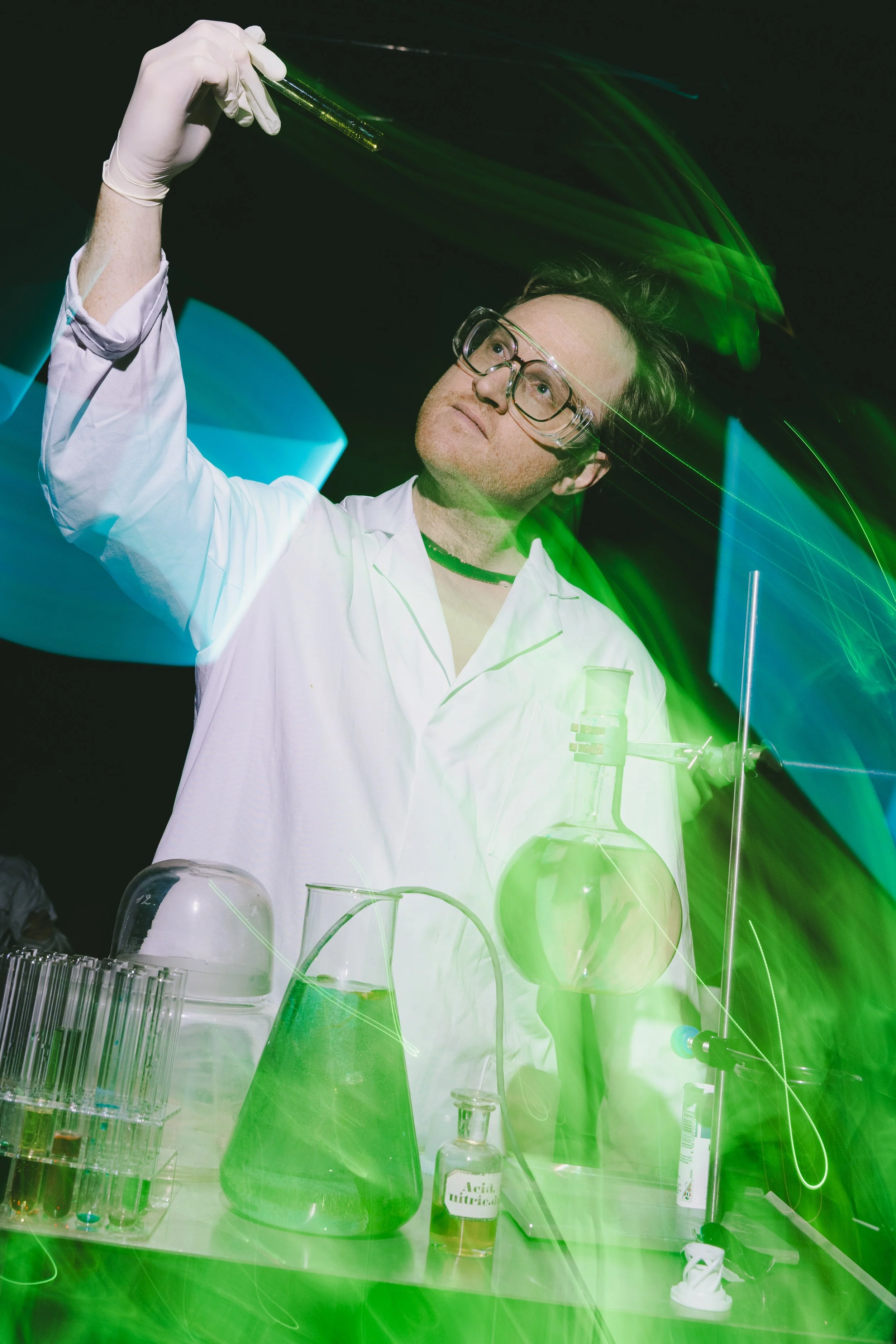Ein Wissenschaftler in Schutzbrille und Laborkittel führt ein Experiment mit chemischen Reagenzien durch, umgeben von Laborgeräten unter grüner und blauer Beleuchtung.