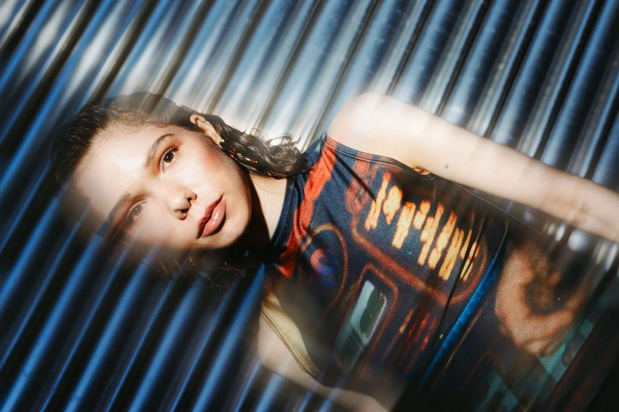 Junge Frau mit dunklem, lockigem Haar und Hoops-Ohrringen sitzt vor einer metallischen, vertikalen Oberfläche, trägt ein T-Shirt mit einem Rockstar-Design, Blick direkt in die Kamera.