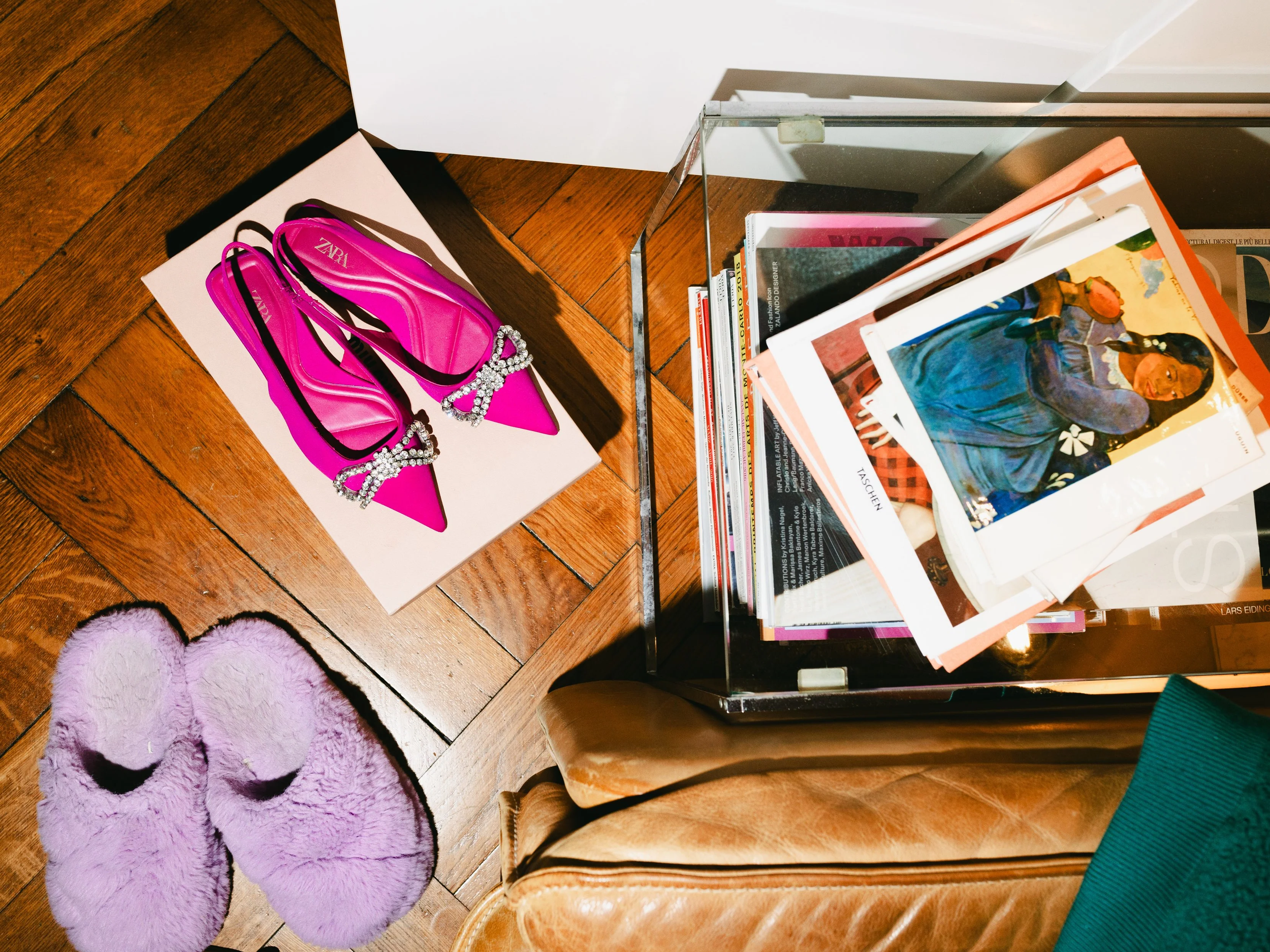 Pinkene Zieratschuhen mit Schleife und Schmuck auf einer weißen Unterlage, lila Kuschelslippers auf einem Holzboden, eine Glasvitrine mit Magazinen und Fotos, ein braunes Sofa mit einem grünen Kissen.