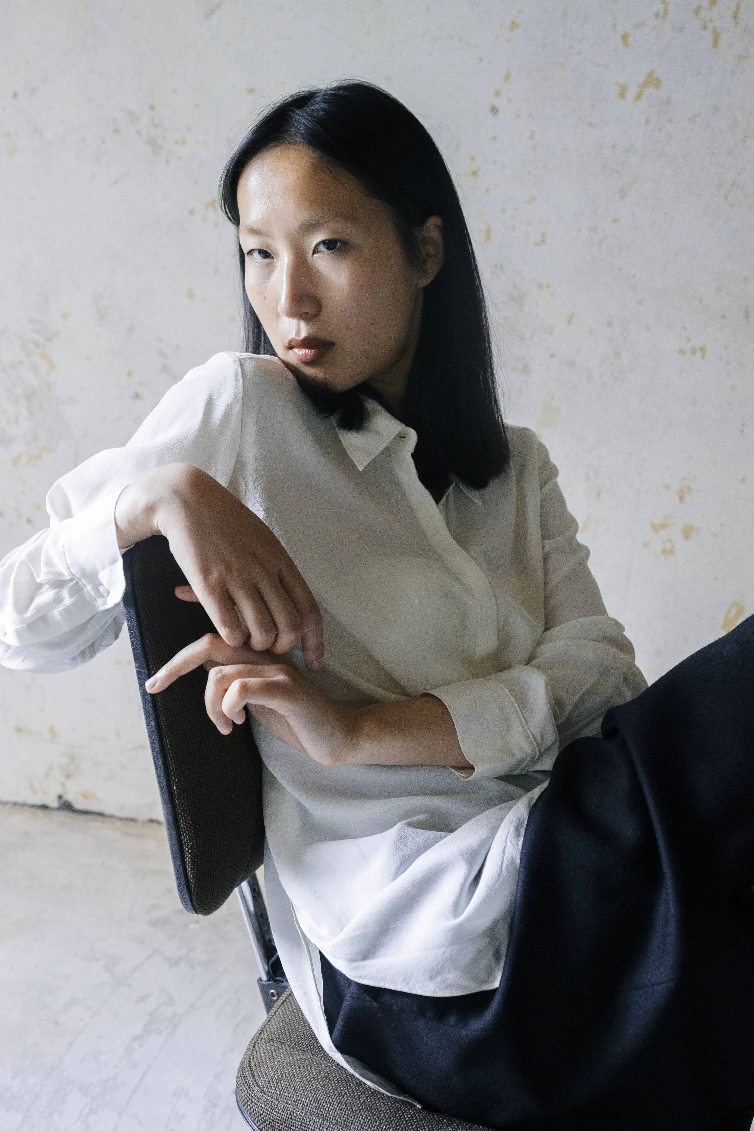Junge Frau mit schwarzen Haaren sitzt auf einem Stuhl vor einer hellen Wand, trägt eine weiße Bluse und blickt nach links.