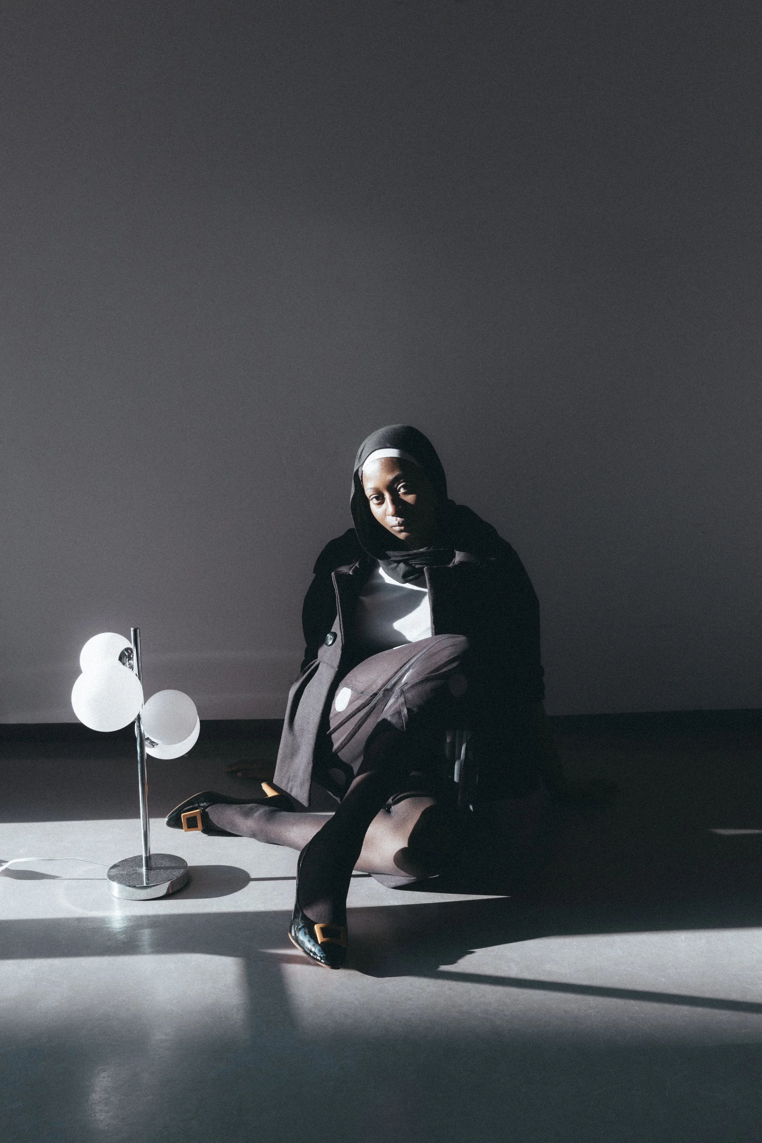 Frau im schwarzen Mantel und schwarzen Schuhen sitzt auf dem Boden neben einem modernen Lampenständer, mit Schatten und diffusen Licht.