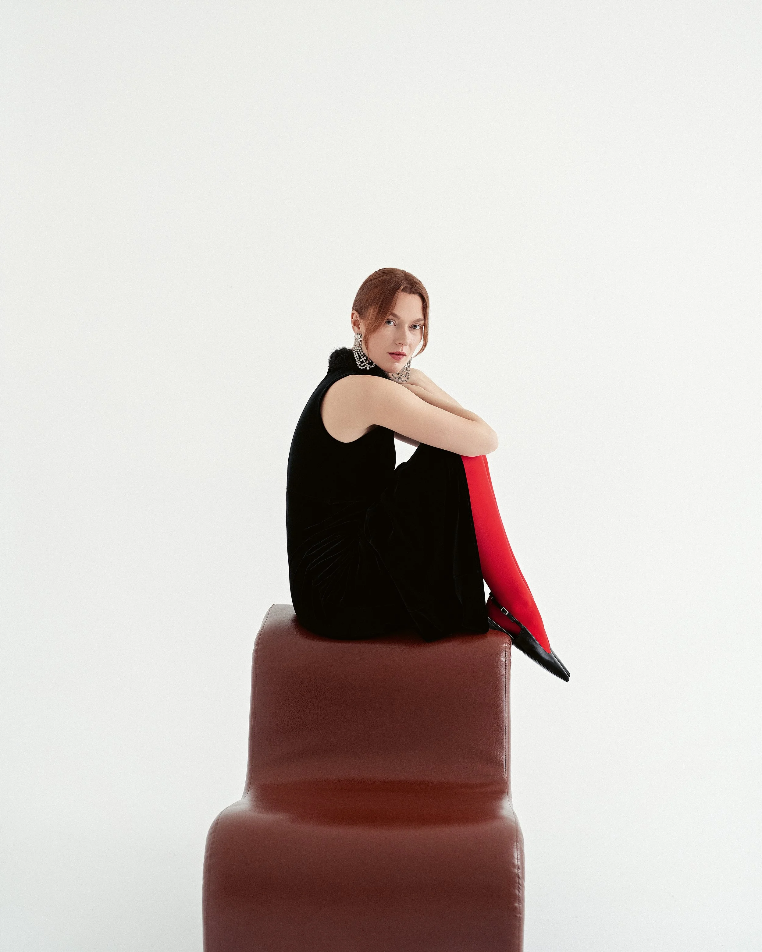 A woman with red hair dressed in a black outfit and red stockings, sitting on a brown chair against a plain white background.
