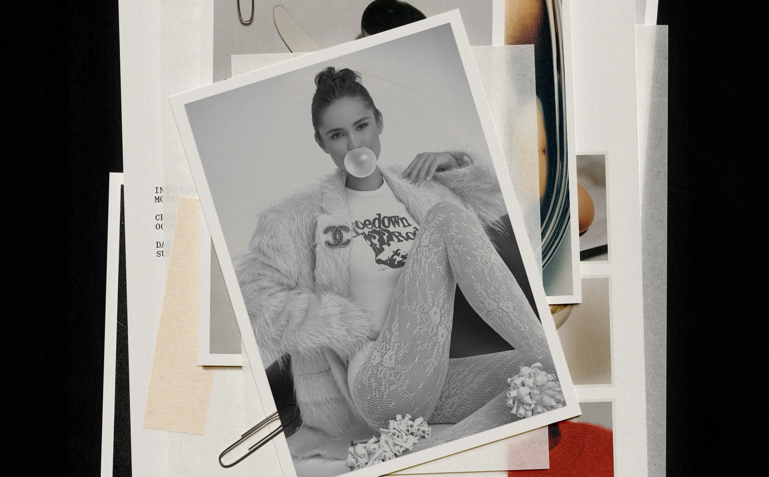 Black and white photo of a young woman sitting on the floor, blowing a bubble with gum, wearing a graphic t-shirt, lace tights, a fur jacket, and sneakers, surrounded by scattered papers and photographs.