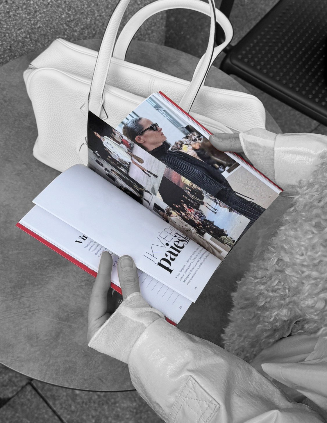 A person wearing gloves is flipping through a fashion magazine, sitting at a table with a white handbag nearby.