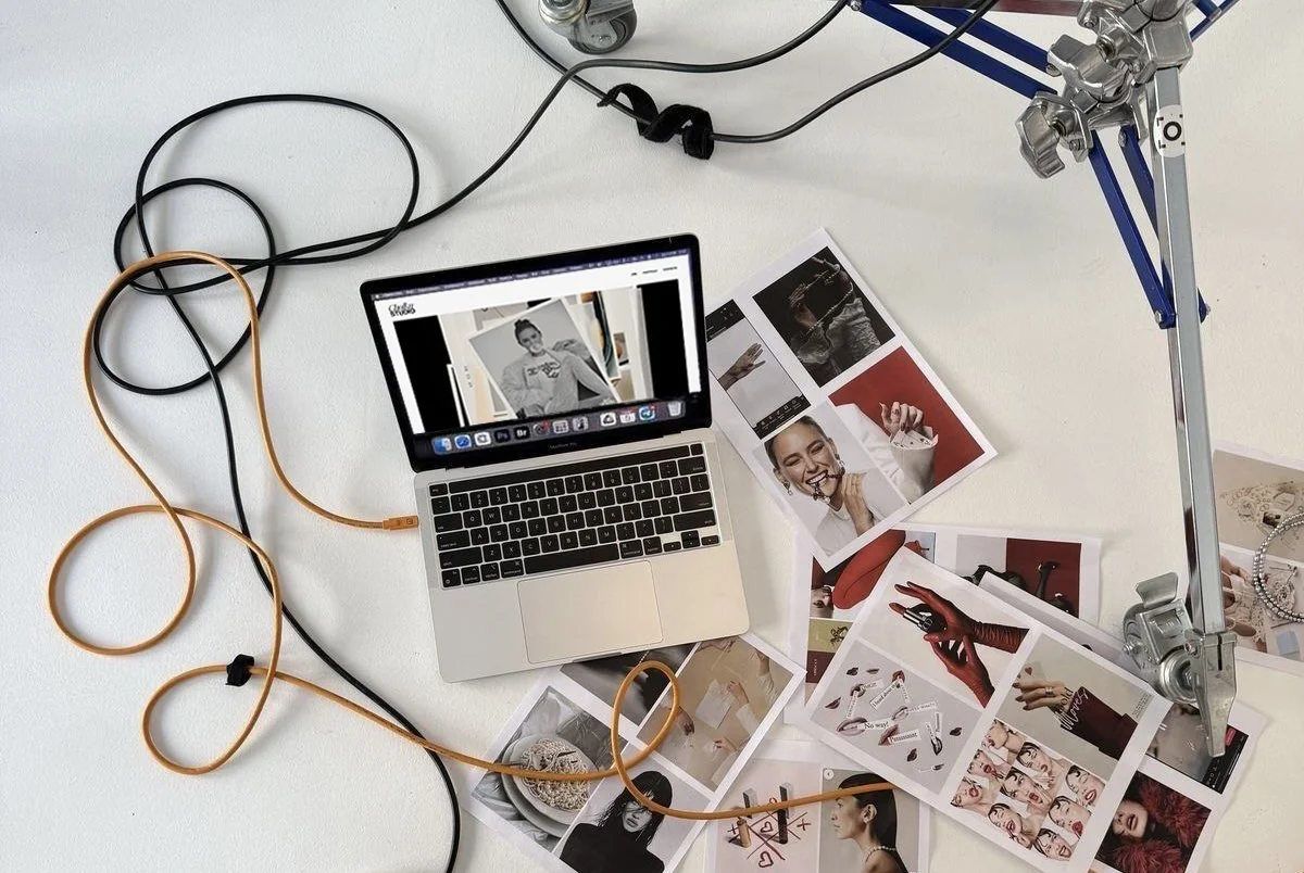 A cluttered desk with a laptop, printed photographs, and various cables, with a camera mounted on an adjustable tripod.