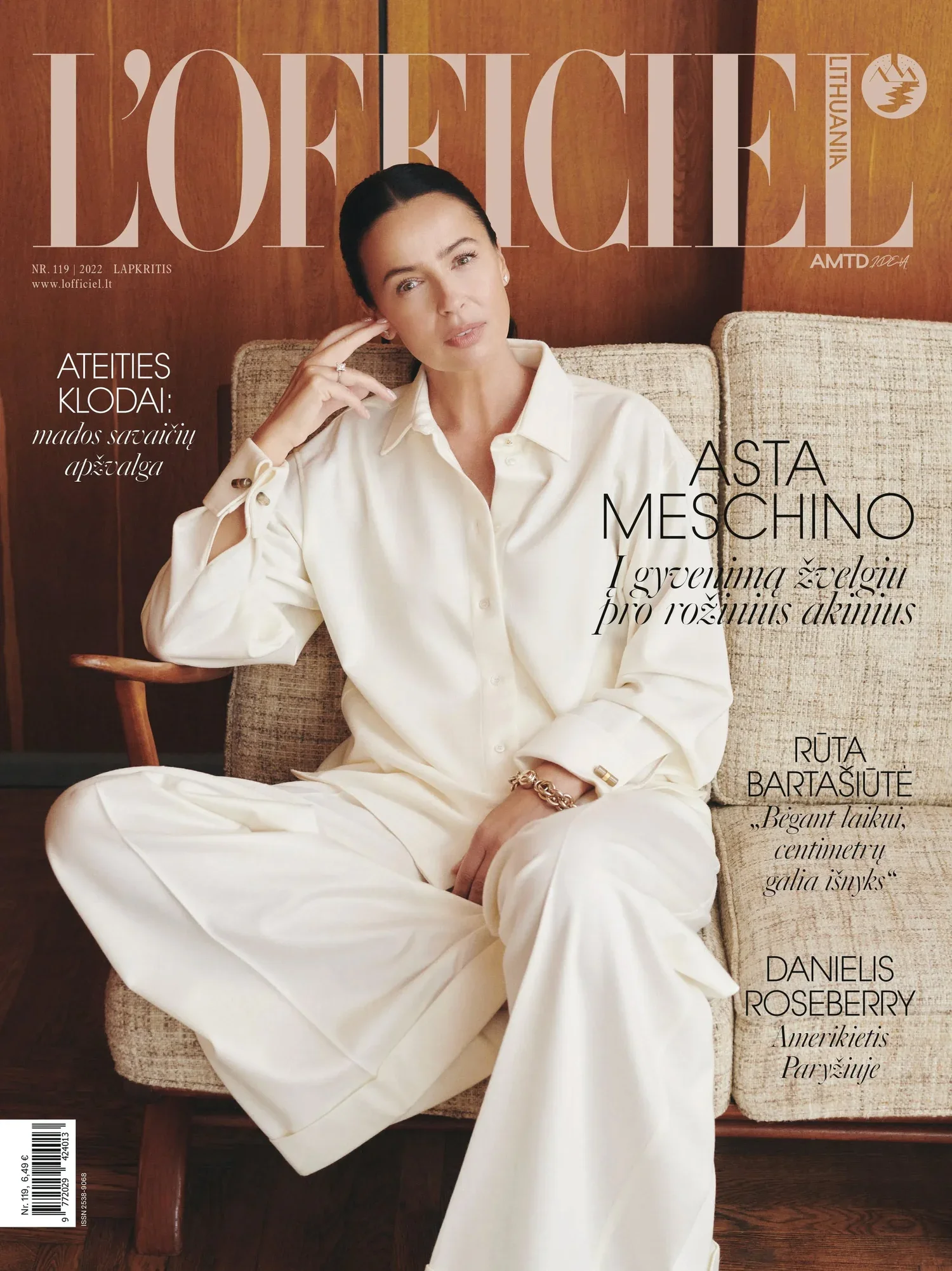 Cover of L'Officiel Lithuania magazine featuring a woman in a cream-colored shirt and wide-leg pants sitting on a beige couch against a wooden wall background.
