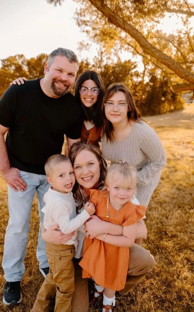 Family of six posing outdoors during golden hour in a park with trees and grass, smiling at the camera.