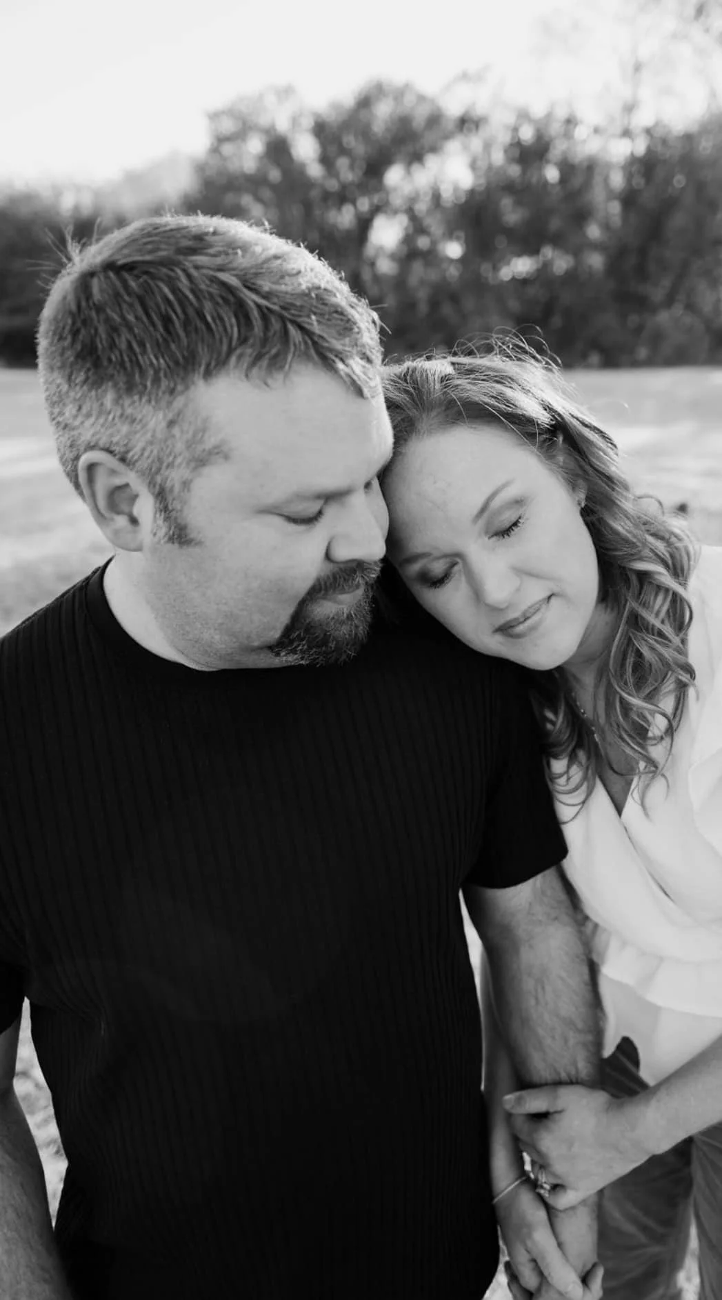 A black-and-white photo of a couple embracing with their heads touching, outdoors with trees in the background.