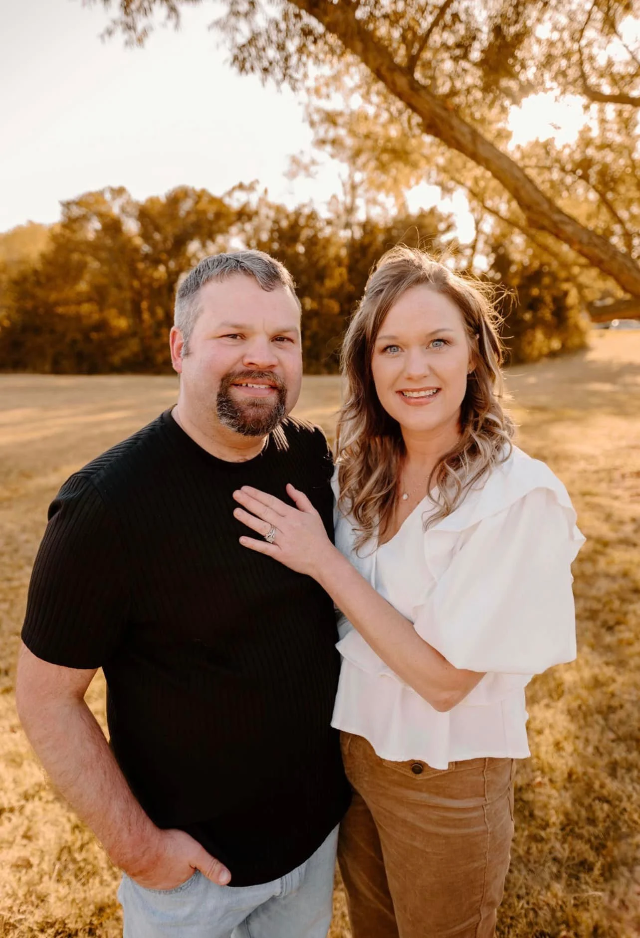 A couple standing outdoors in a park during golden hour, smiling at the camera with a large tree in the background. The woman is wearing a white top and the man is in a black shirt, and she has her hand with a ring on her chest.
