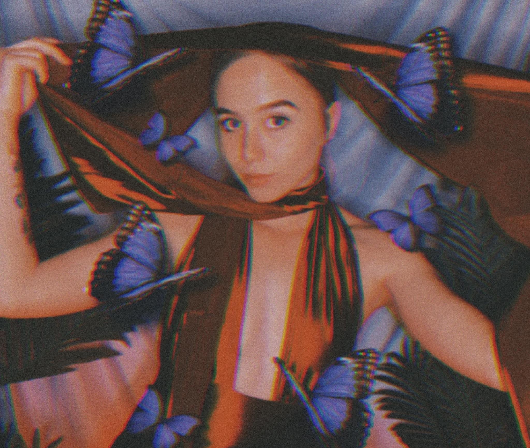 A woman with long dark hair and light skin lying on a blue bedspread, wearing a blue shirt. She is holding a large butterfly-shaped object that covers her head and shoulders, with the butterfly's wings spread around her.