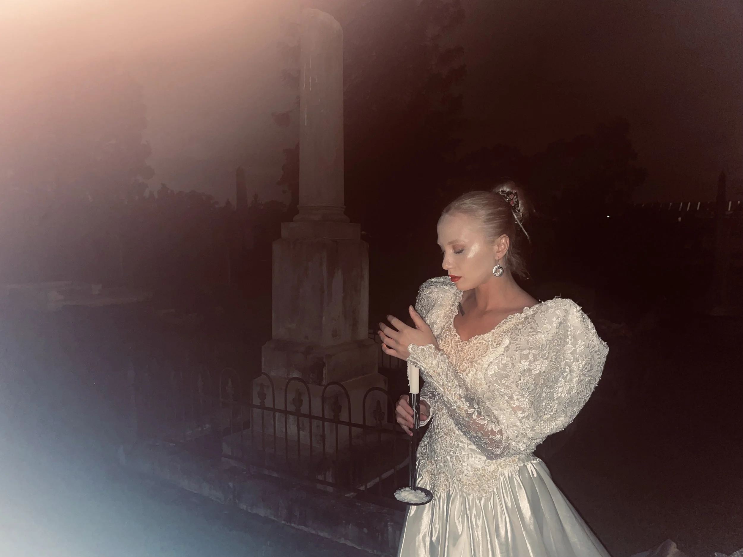 A woman dressed in a wedding gown, holding a candle and standing outdoors at night near a small fence, with trees and a tall monument visible in the background.