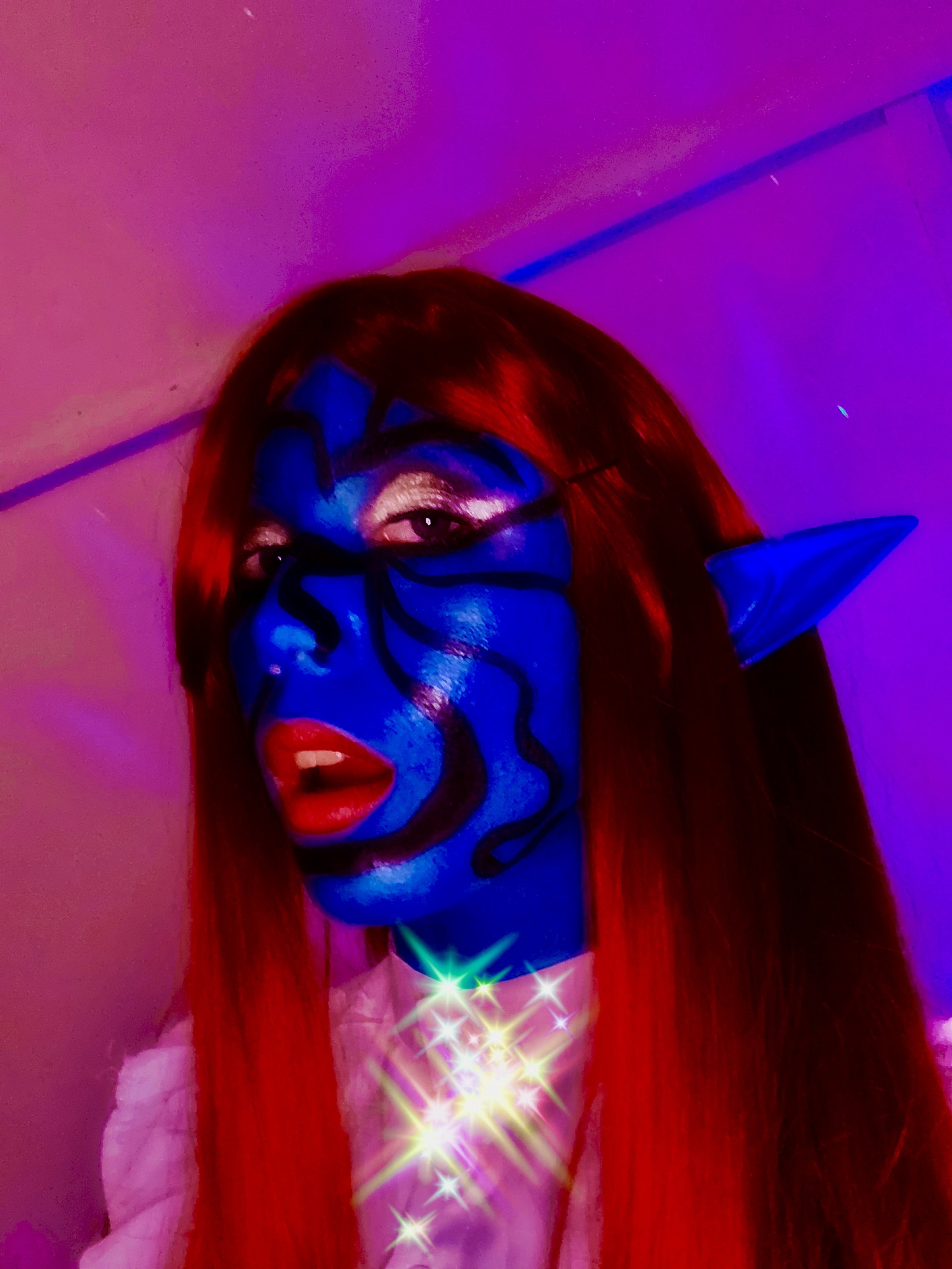A person with red hair and blue face paint, wearing glasses and elf ears, lit by purple and pink lighting and surrounded by glowing stars.