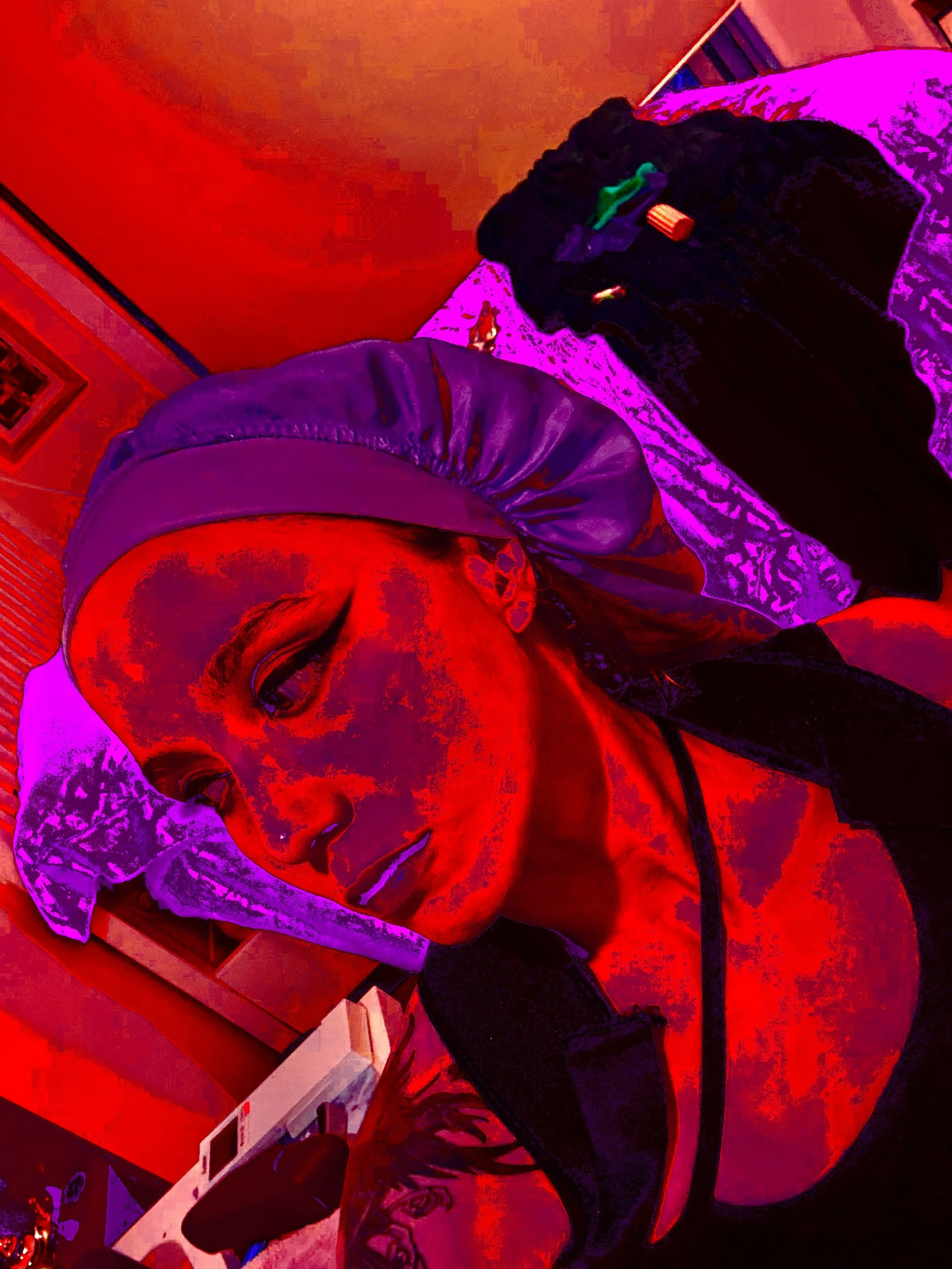 A young man with a tattooed arm is taking a selfie. He is wearing a dark jacket and a baseball cap and is in a room with reddish and purple lighting.
