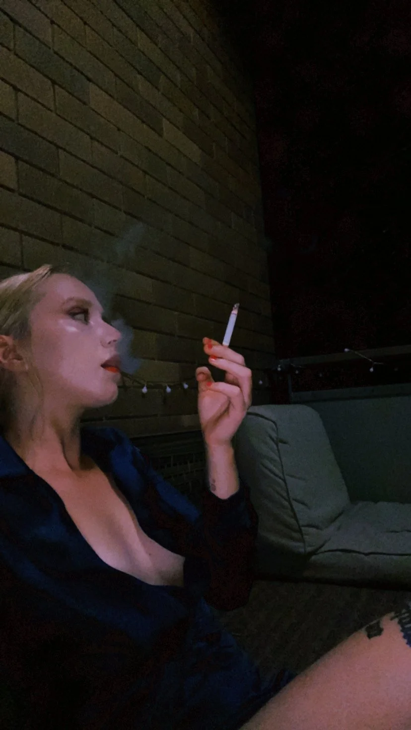 A woman with blonde hair and makeup, sitting outside at night against a brick wall, smoking a cigarette, with a tattoo on her thigh, and a cushion nearby.