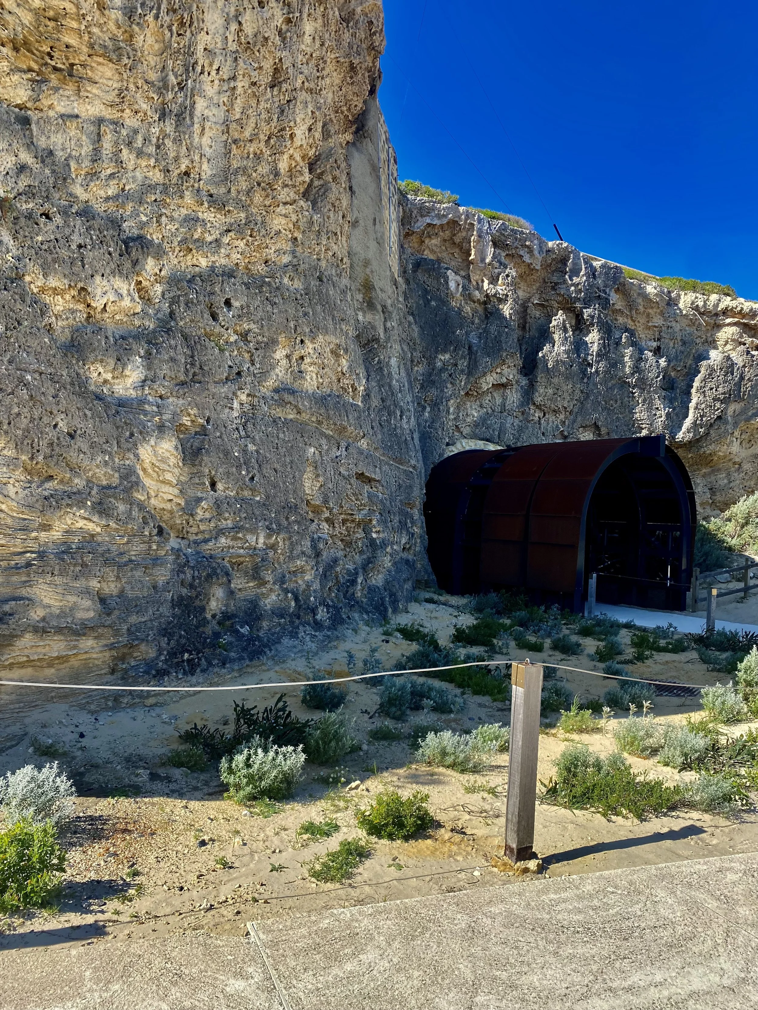 A large natural rock cliff with a dark tunnel entrance at the base, surrounded by desert plants and a sandy area with a wooden post and rope barrier in the foreground under a clear blue sky.