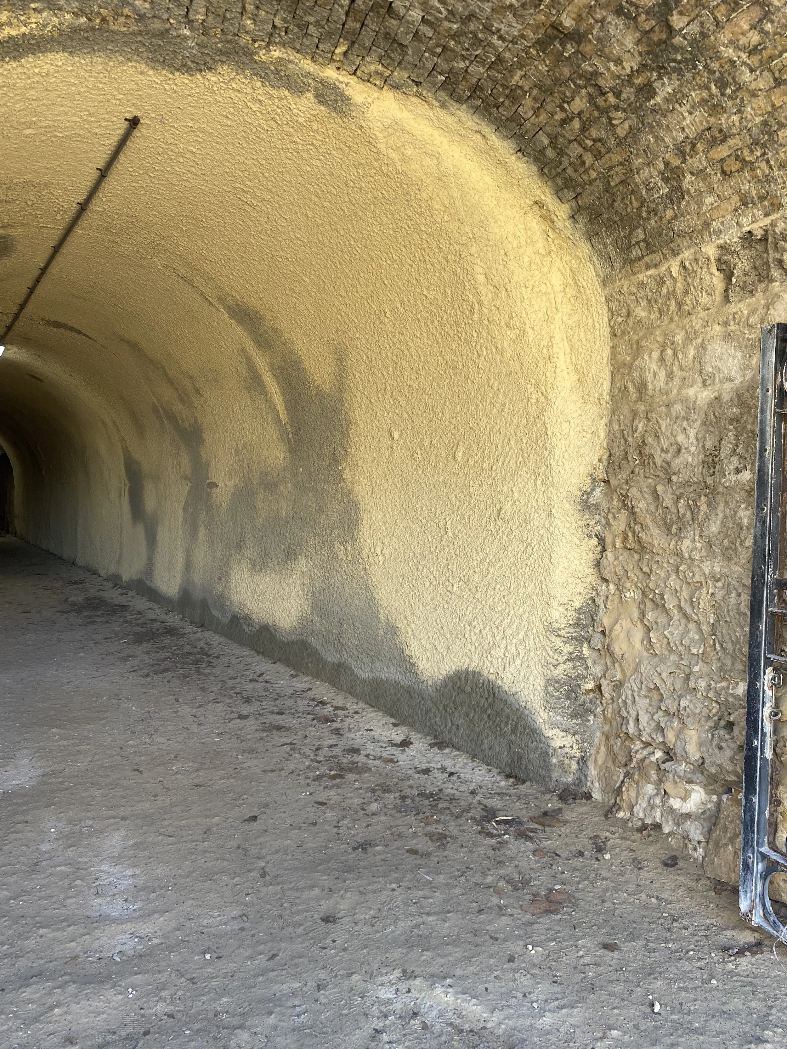 The image shows the interior of a tunnel with textured yellow-painted ceiling and walls made of brick and stone. The ground is dirt, and there is a metal structure on the right side of the image.