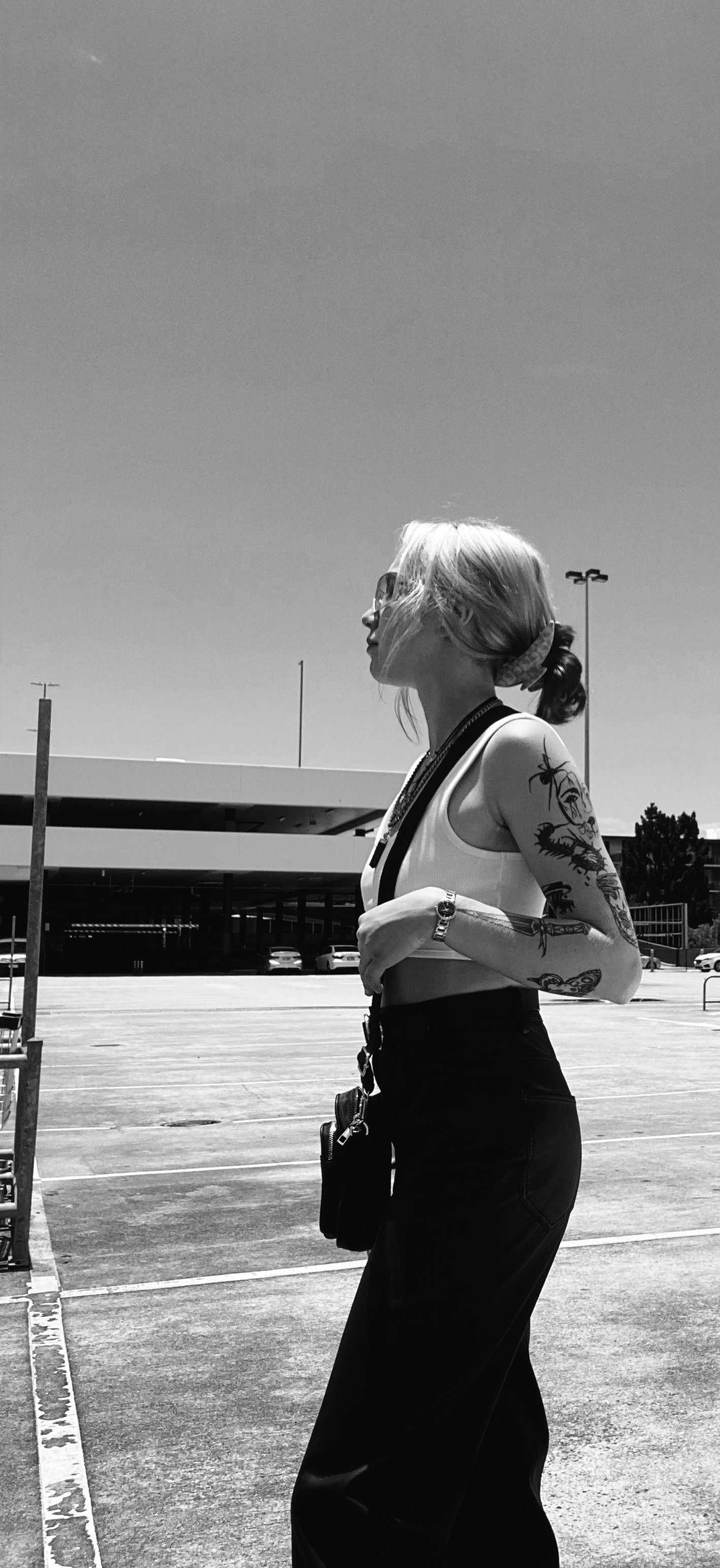 Side view of a woman with blonde hair tied back, wearing sunglasses, a white crop top, and black pants, standing in a parking lot with cars and a building in the background on a clear day.