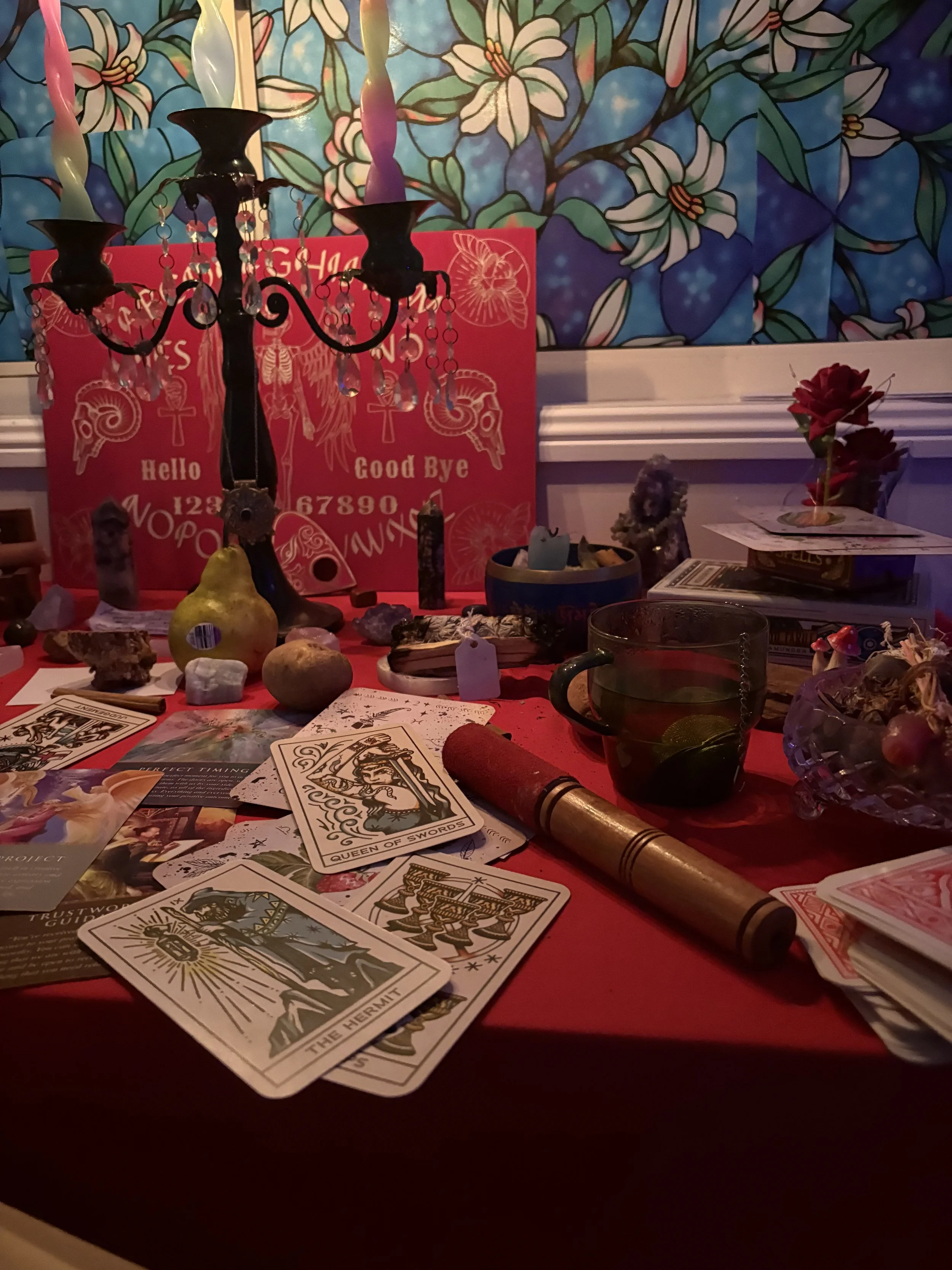 A table covered with various objects including tarot cards, crystals, a small cauldron, and a candle holder with unlit candles, with a colorful floral stained glass window in the background.