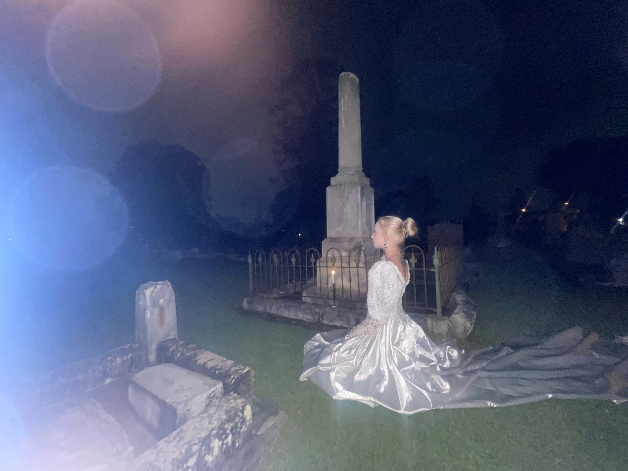 A woman dressed in a silver gown standing in front of a monument at night. The monument is tall with a column structure, illuminated in the dark sky. There is a fence surrounding the monument, rocks, and grass at its base.