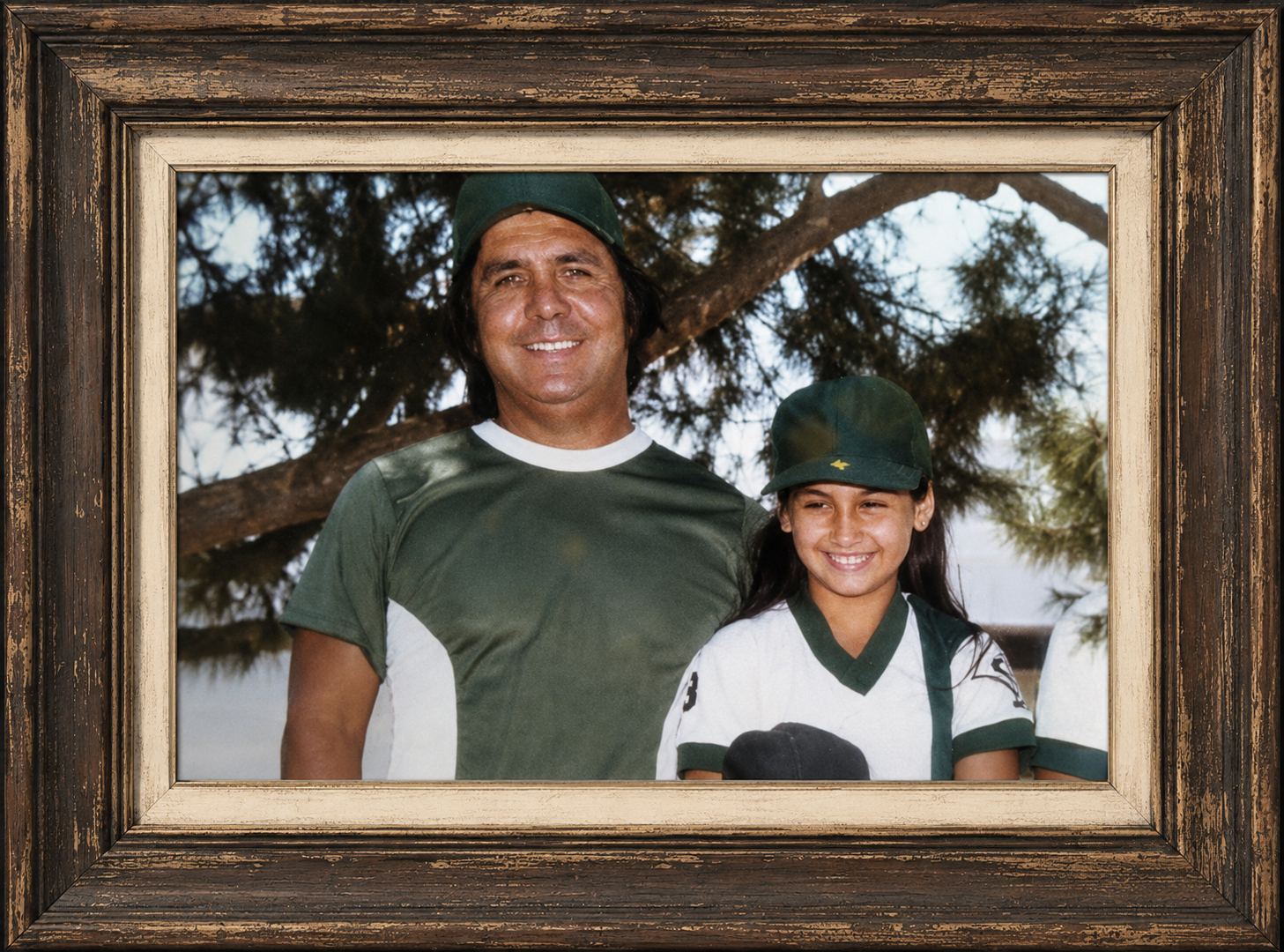 A framed photograph of a man and a young girl outdoors, both smiling, with trees in the background. The man and girl are wearing green hats and athletic shirts.