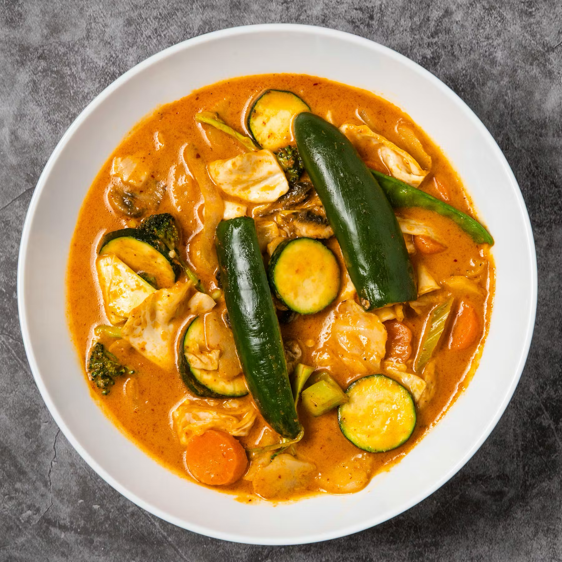 A bowl of vegetable curry with zucchini, jalapeño, broccoli, carrots, and cabbage in an orange sauce.