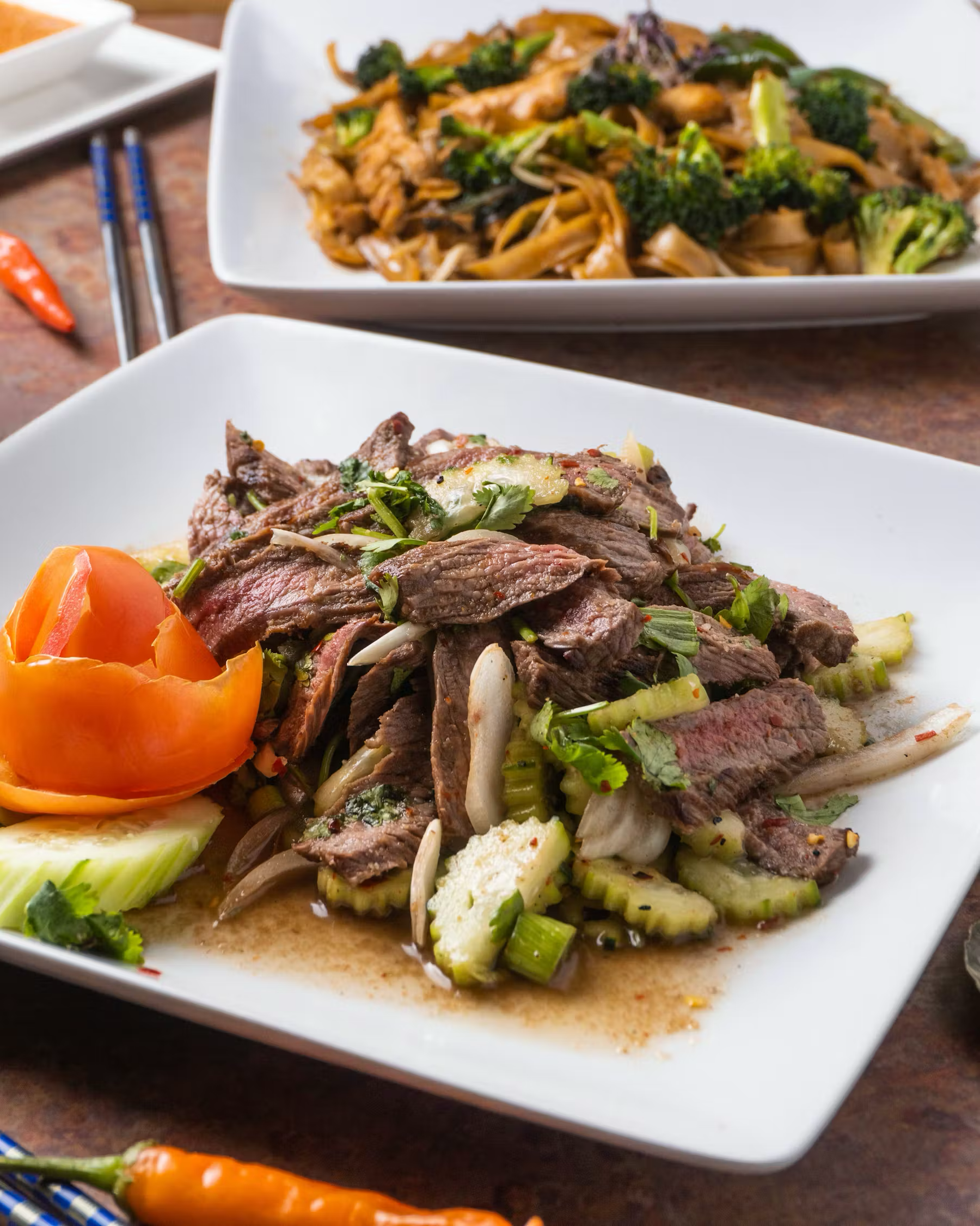 A plate of sliced beef with herbs and vegetables, garnished with a tomato and cucumbers, on a white square plate. In the background, chopsticks and a dish of stir-fried noodles with broccoli.
