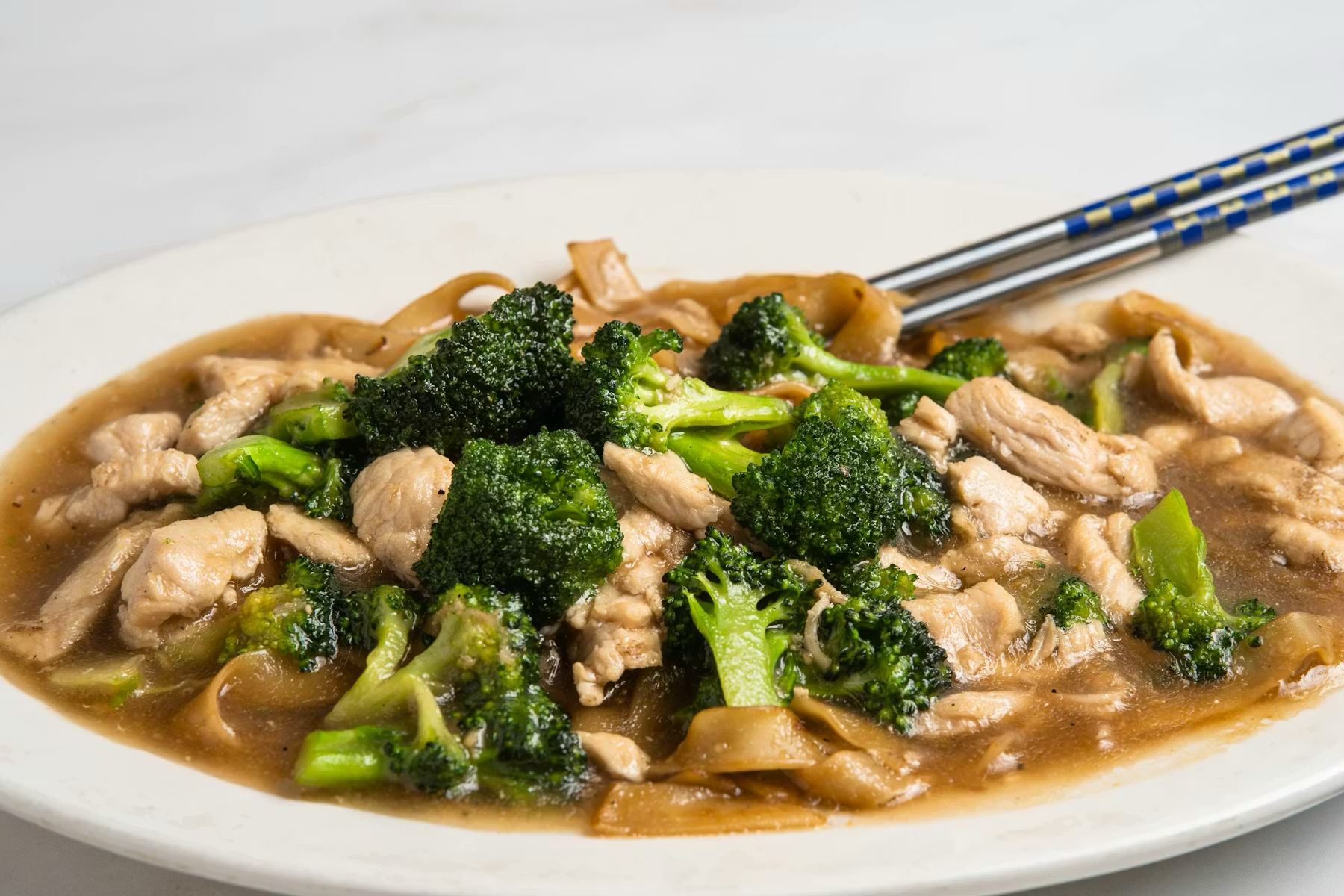 Stir-fried broccoli, chicken, and noodles in sauce on white plate with chopsticks.