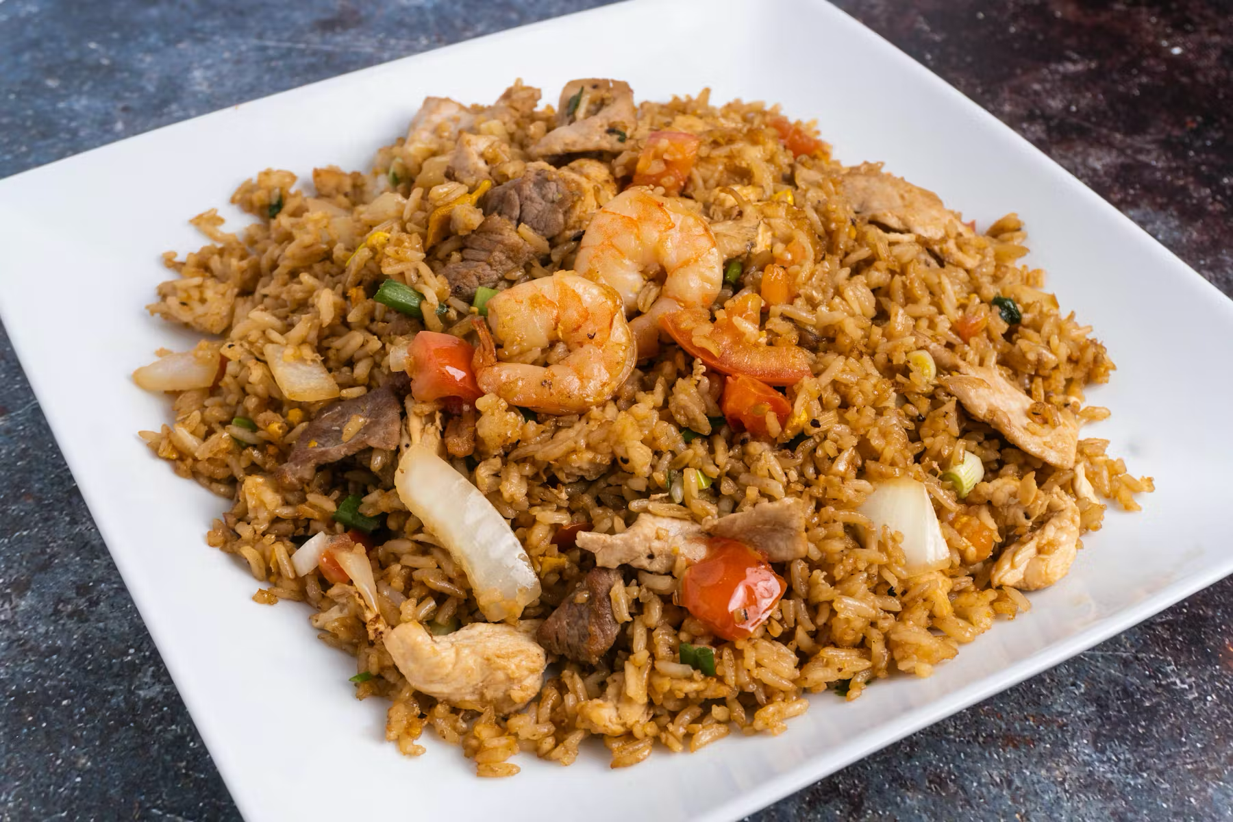 Plate of fried rice with shrimp, chicken, beef, and vegetables, on a dark surface.
