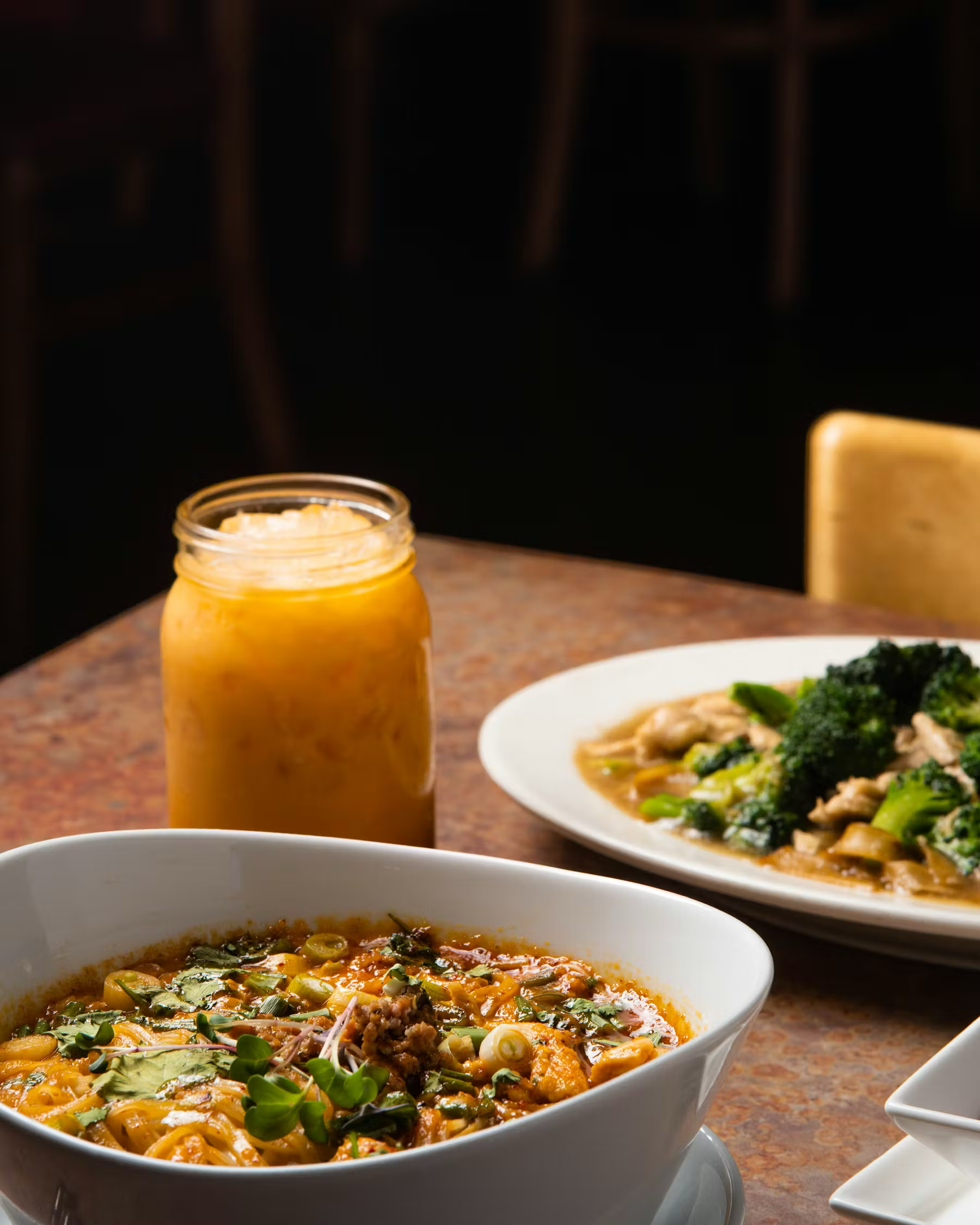 A bowl of Asian noodle soup with vegetables and herbs on a table with a jar of mustard and a plate of broccoli and chicken in the background.