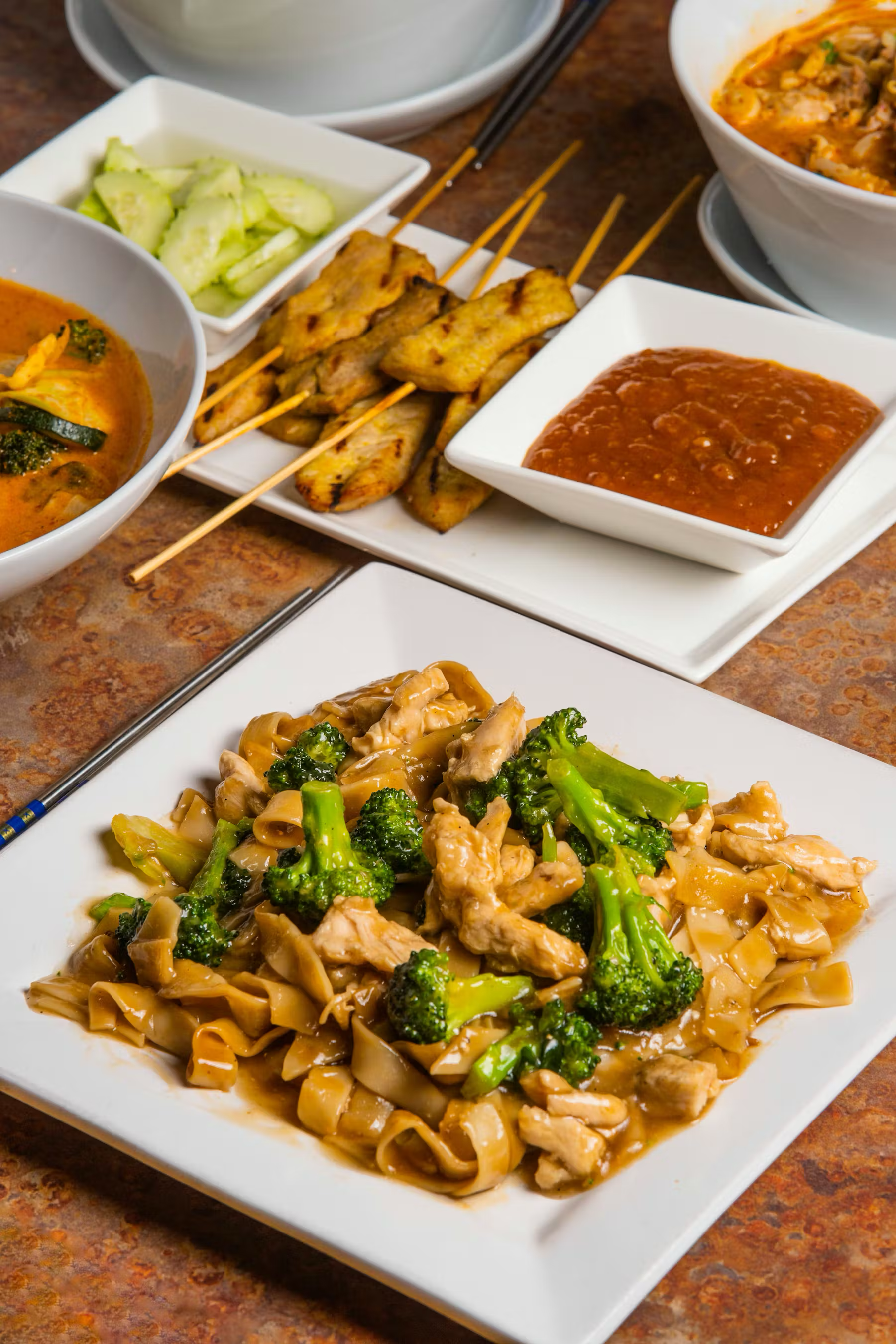 Plate of stir-fried chicken with broccoli and noodles, with side dishes including skewered grilled chicken, cucumber slices, a bowl of curry, and a bowl of chili.
