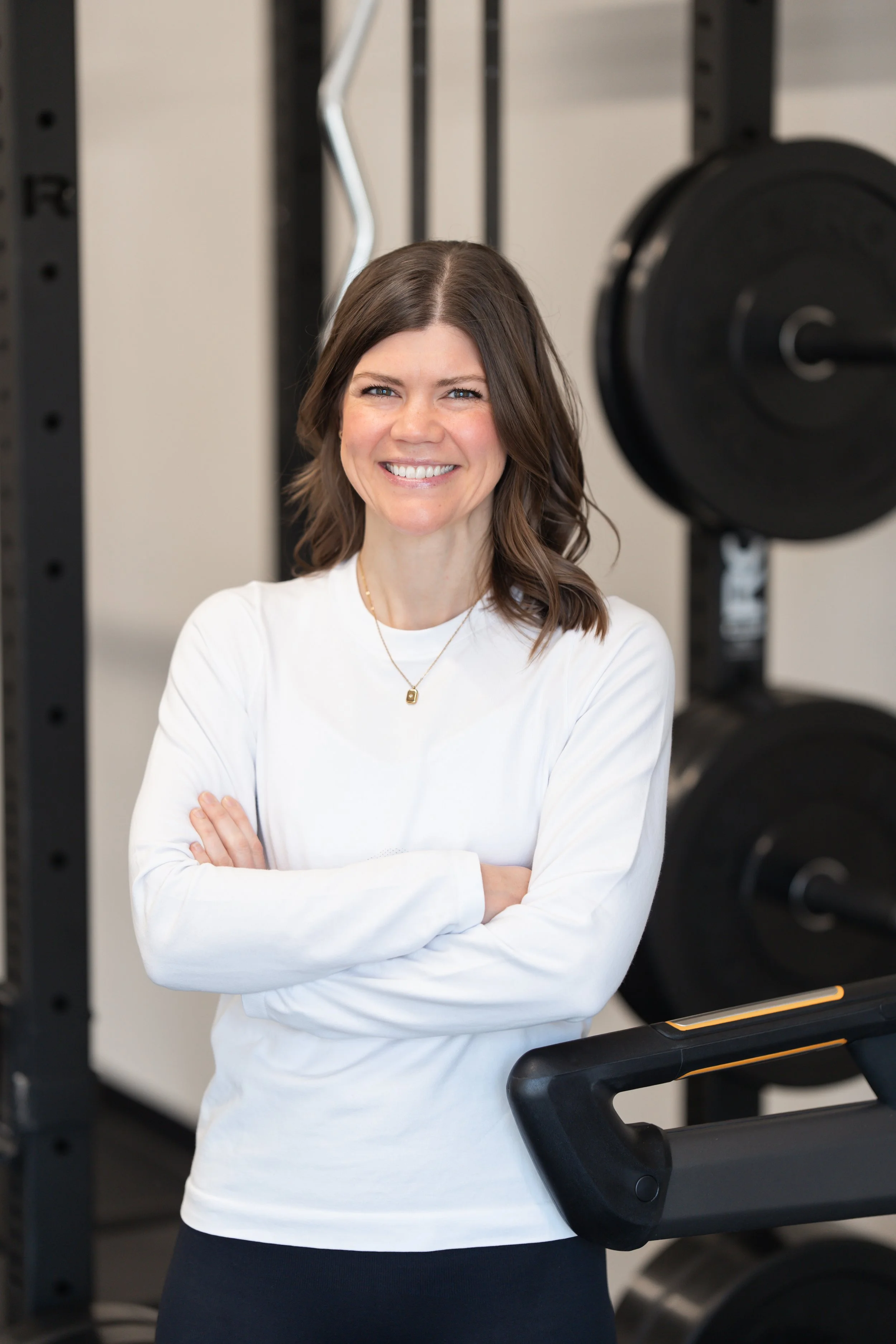 A smiling woman with shoulder-length brown hair, wearing a white long-sleeve shirt, standing with arms crossed in front of gym equipment and weight plates.