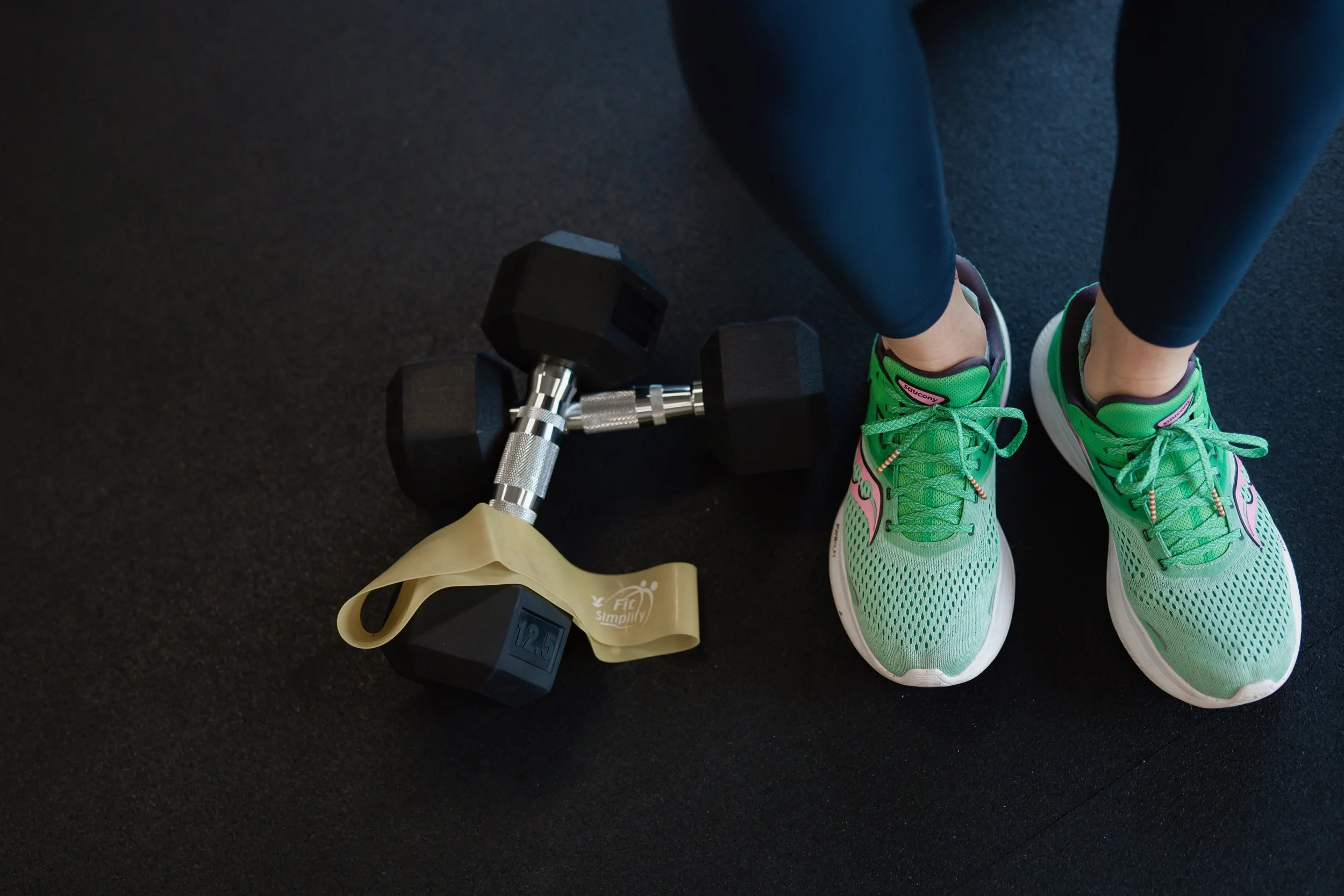 A pair of bright green athletic shoes worn by a person sitting on a black gym floor, with two black and silver dumbbells nearby.