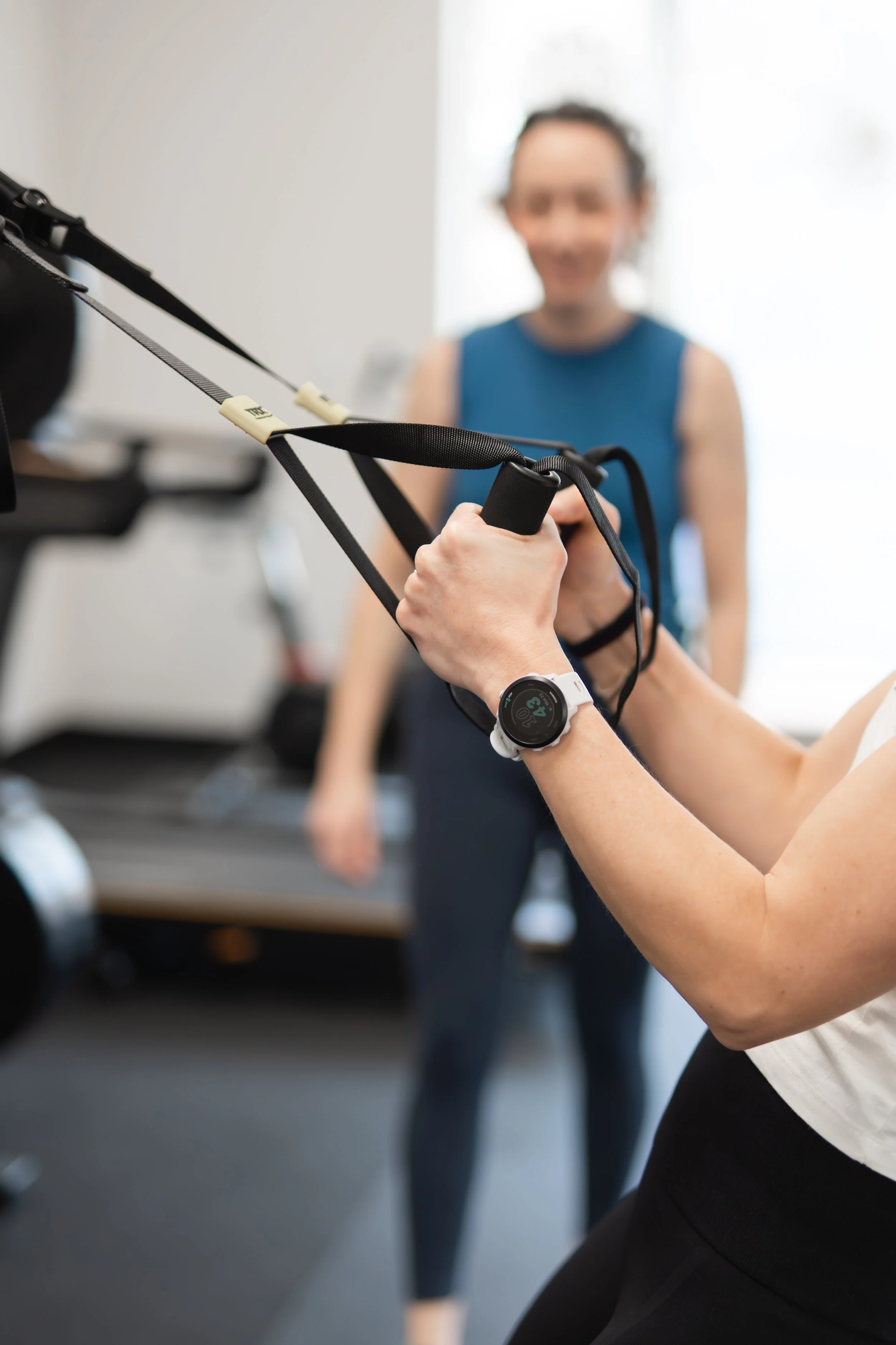 Person exercising with resistance bands while wearing a smartwatch, with another person observing in the background.