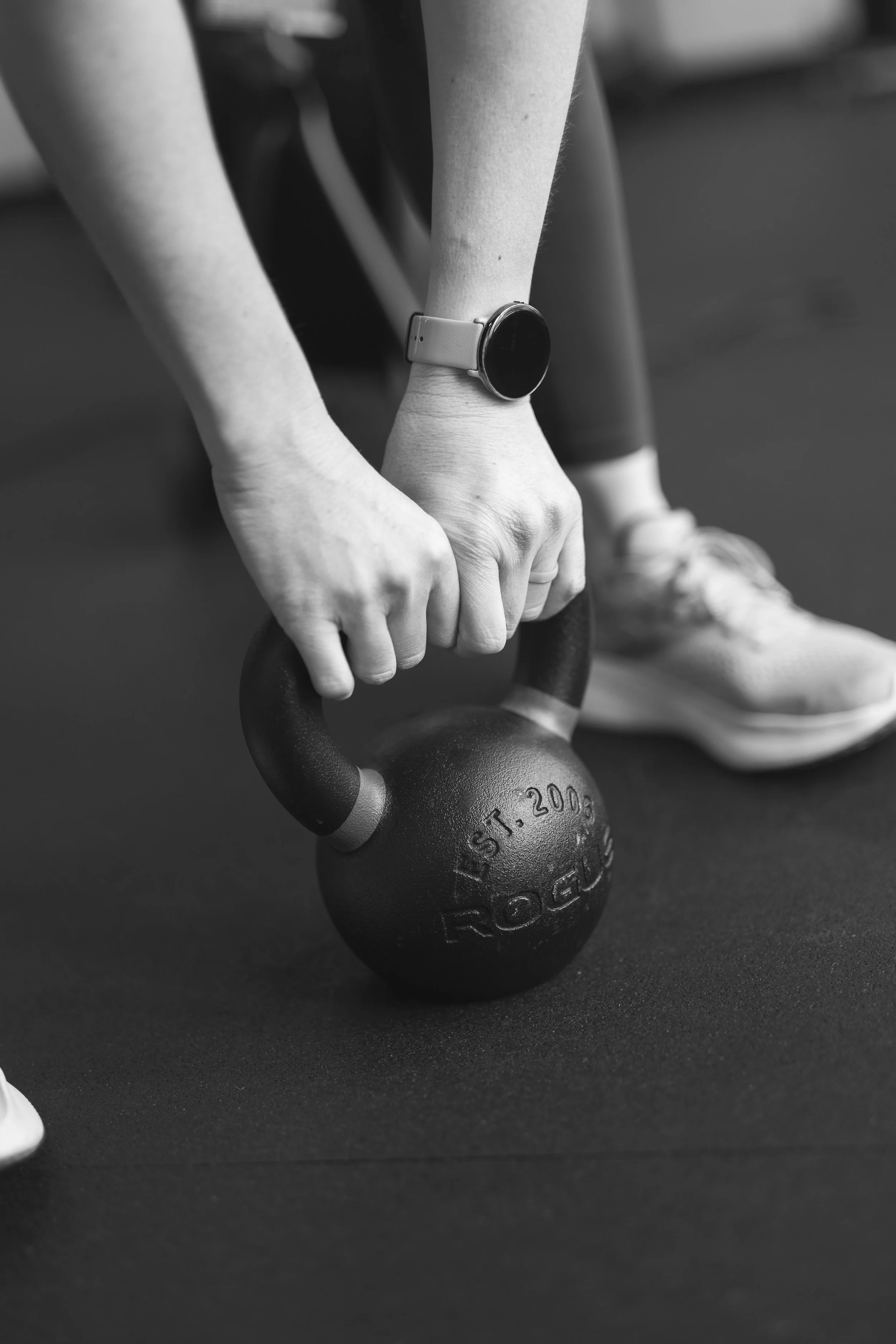 A person wearing sneakers and a smartwatch is holding a kettlebell on a gym floor.