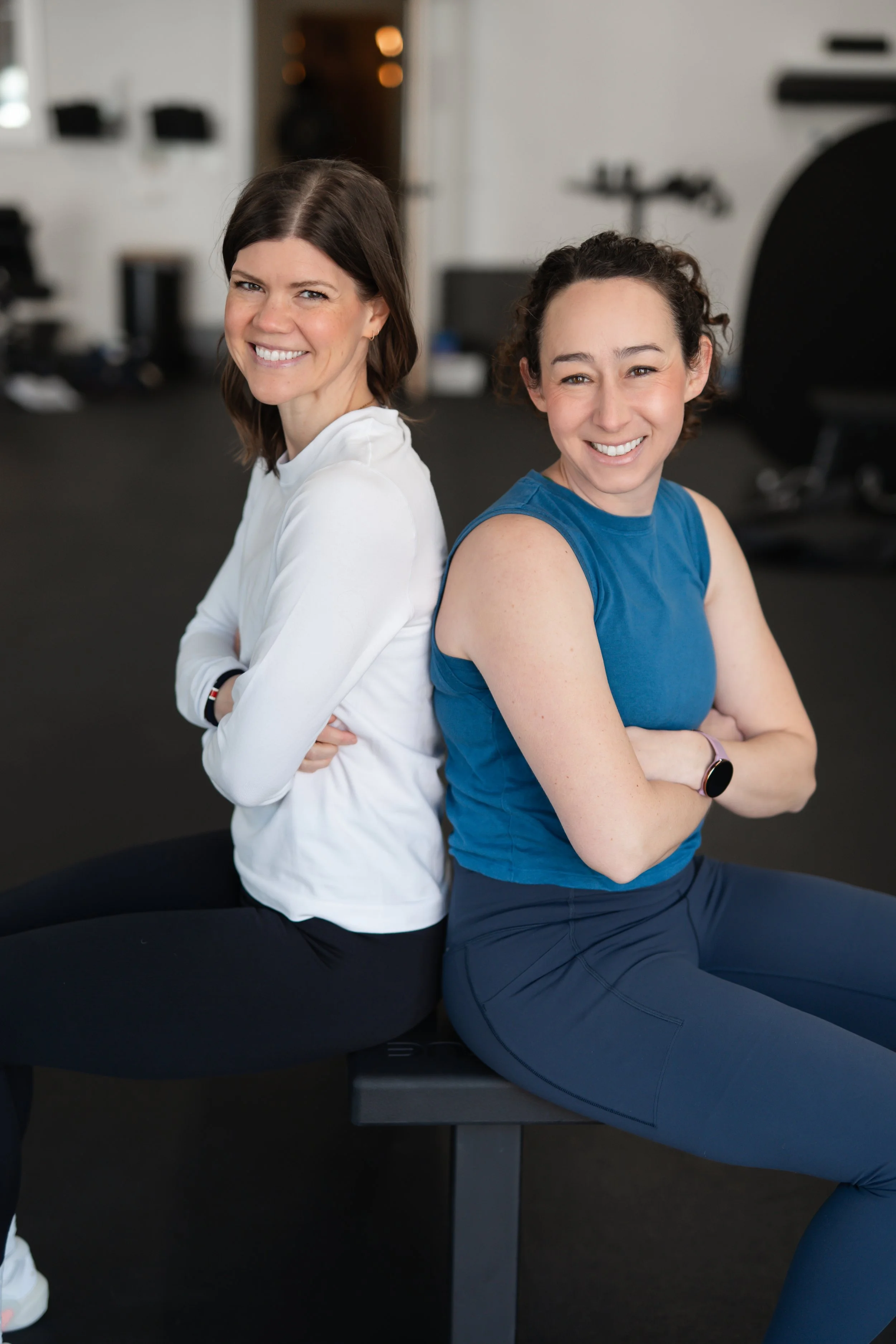 Two women sitting back-to-back on a gym bench, smiling and crossing their arms.