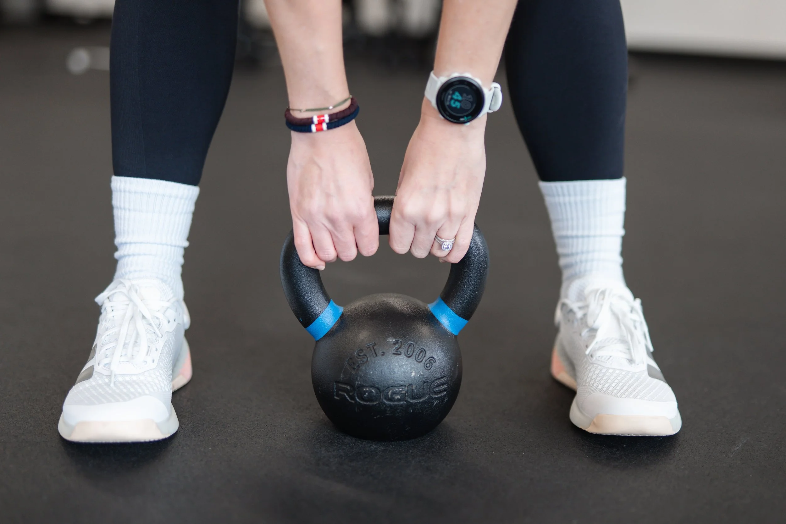 Person wearing white sneakers and socks, bending down to grip a black kettlebell with blue markings in a gym with a black floor.