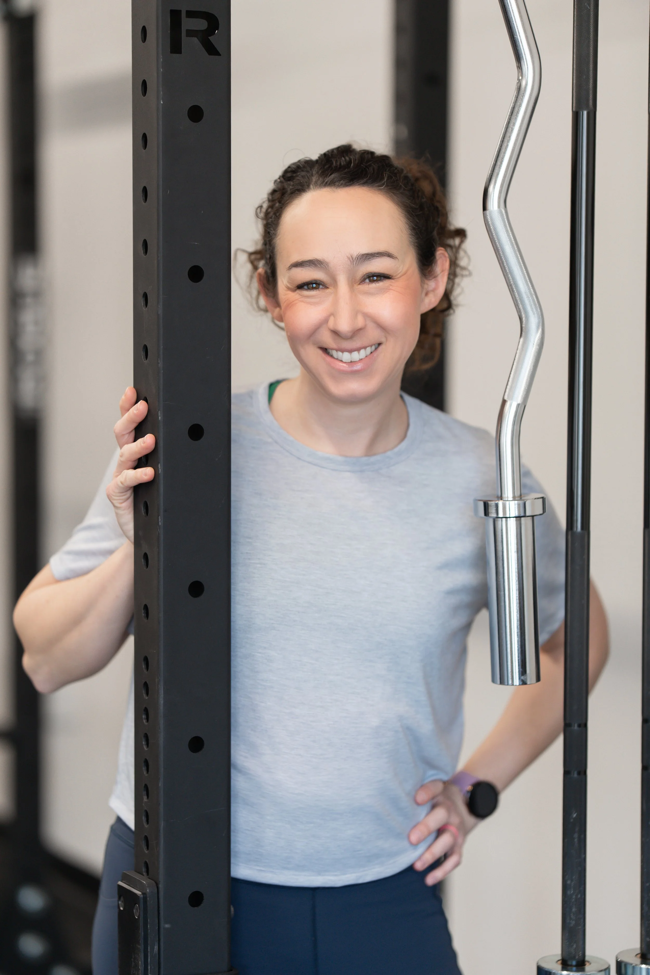Smiling woman standing in a gym, holding a vertical black fitness rack with one hand and resting her other hand on her hip, wearing a gray shirt and a black smartwatch.